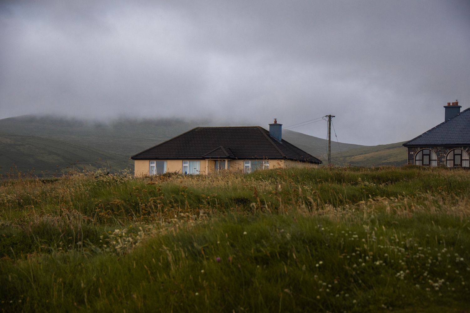 Rural landscape study. Focus on the low, geometric cottage nestled within the vast, misty hills. Atmospheric exploration of domestic scale.