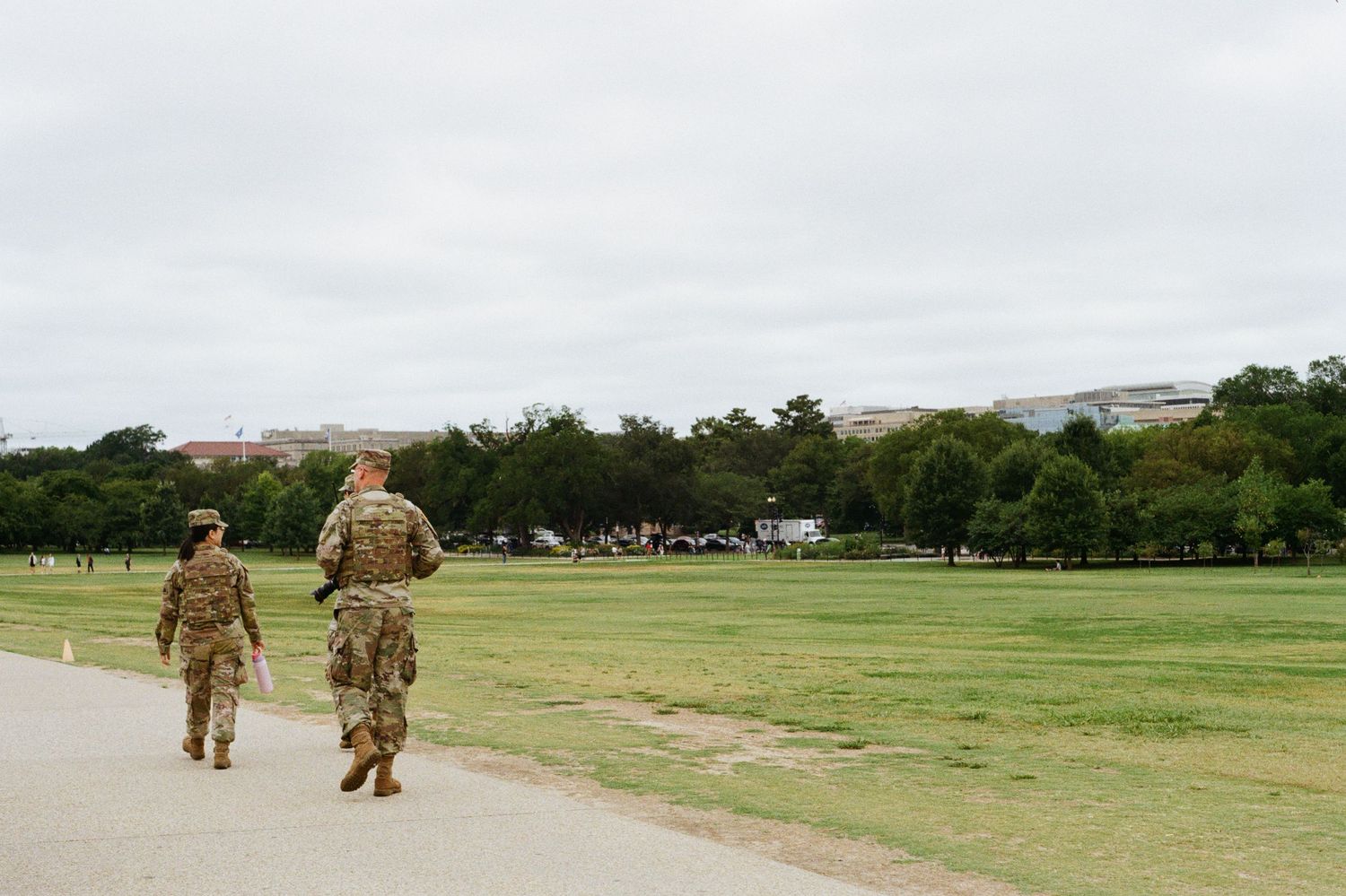 National Guard walk across the vast green lawns of the National Mall in Washington, D.C., under a soft, overcast summer sky.