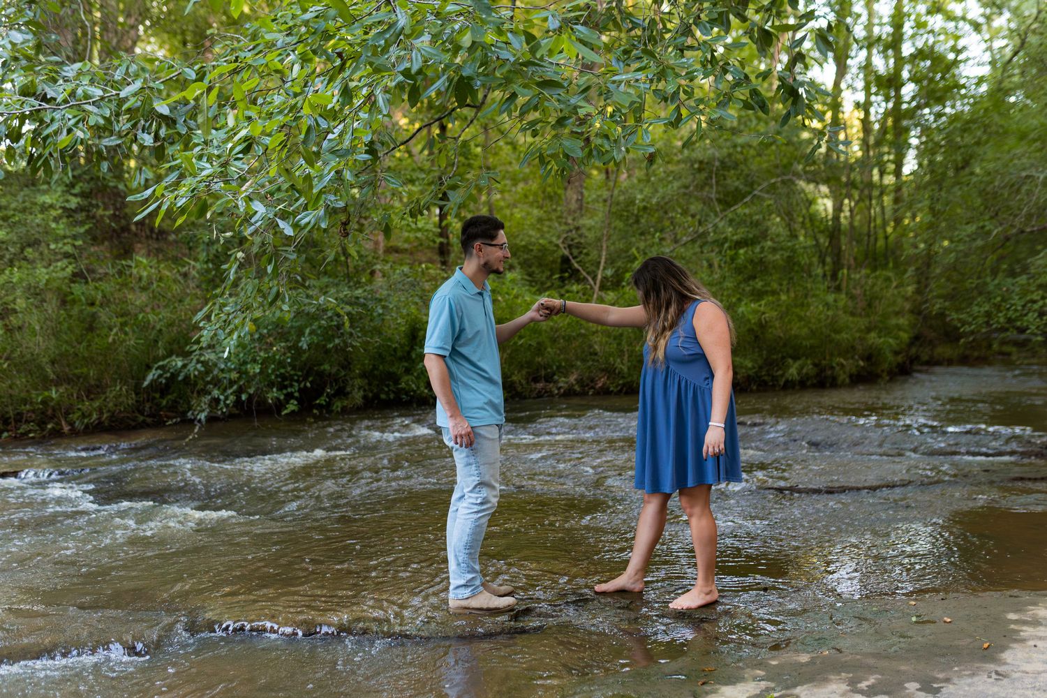 Two people explore a shallow creek bed surrounded by lush greenery on a summer day.