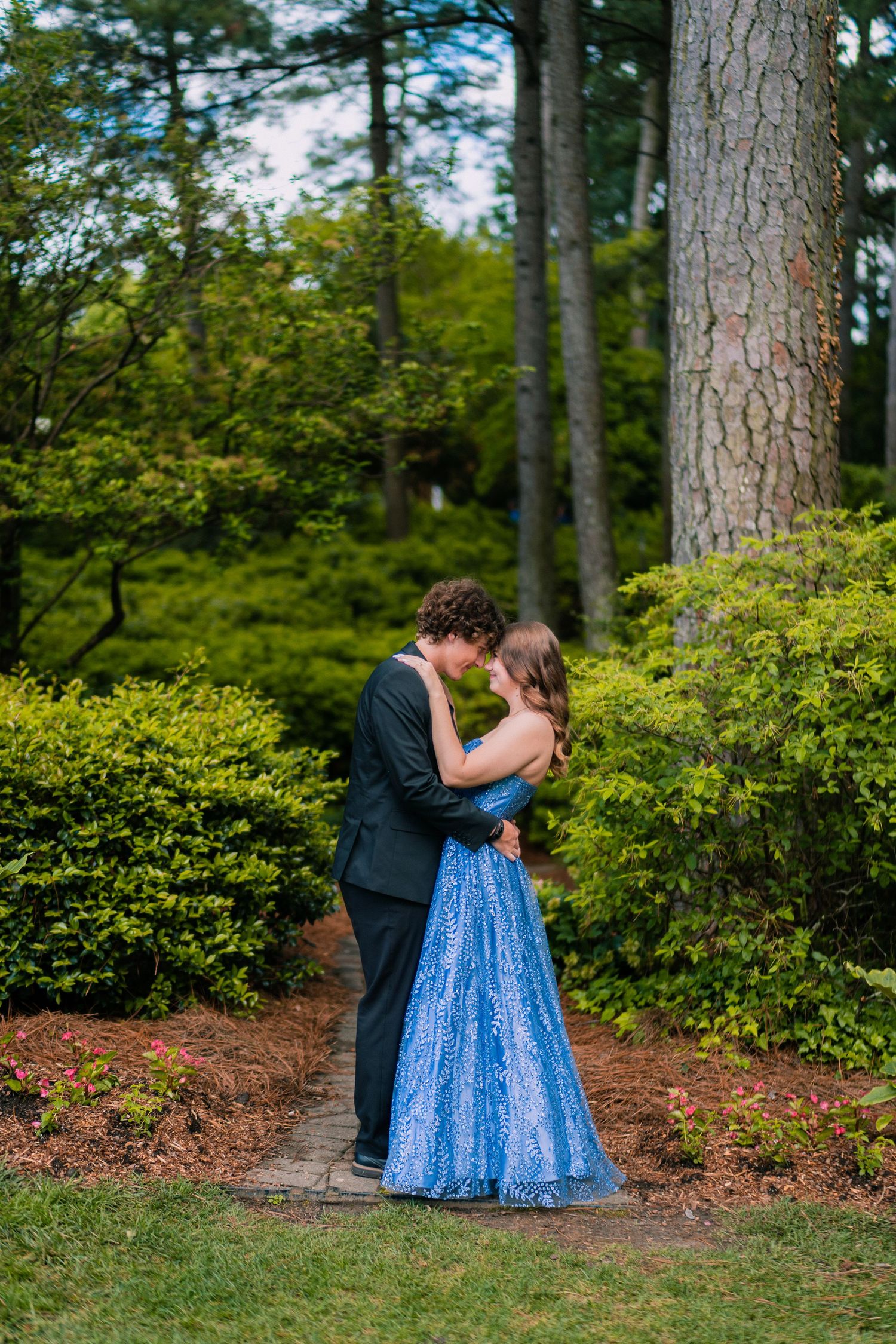 A couple in formal attire embracing in a lush garden setting with tall pine trees.