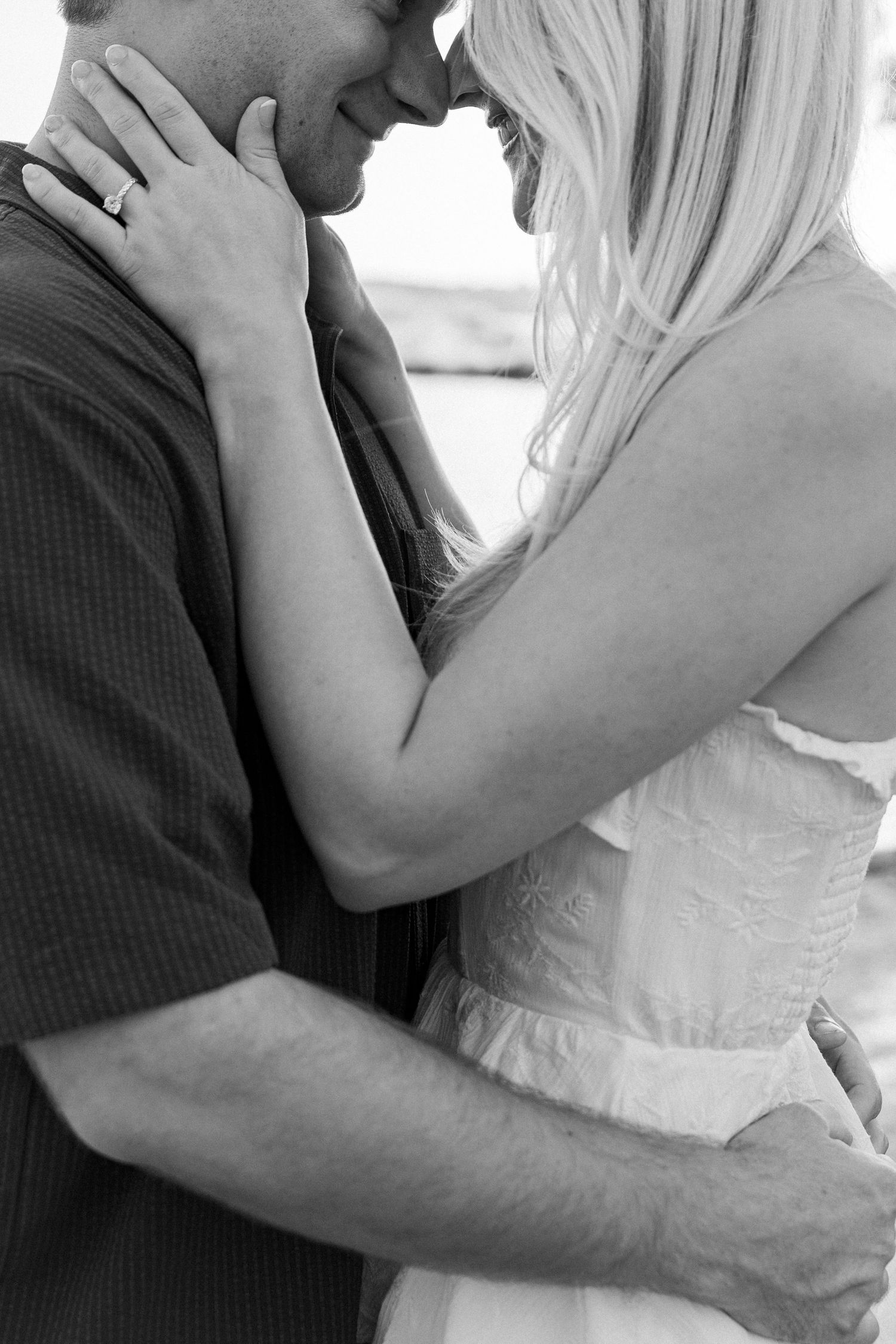 Black and white photo of a romantic couple embracing closely, woman in white dress with engagement ring visible.