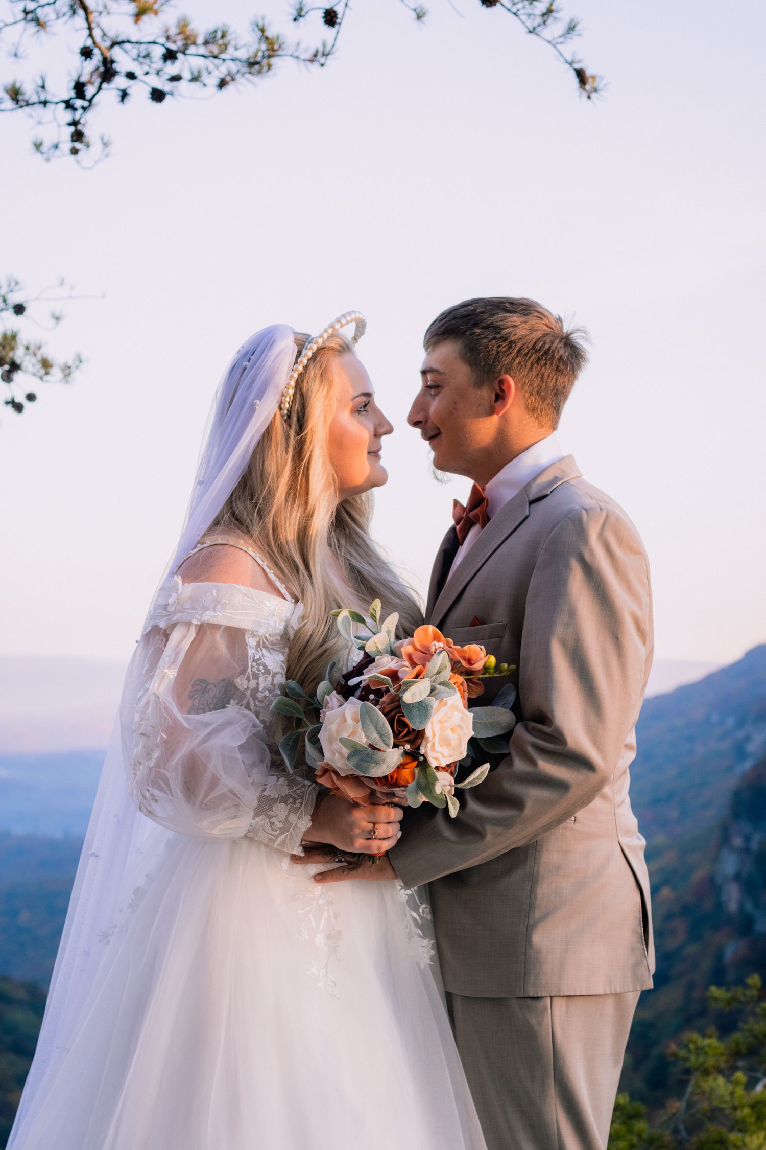 A couple in wedding attire share an intimate moment on a mountaintop with a scenic mountain landscape behind them.