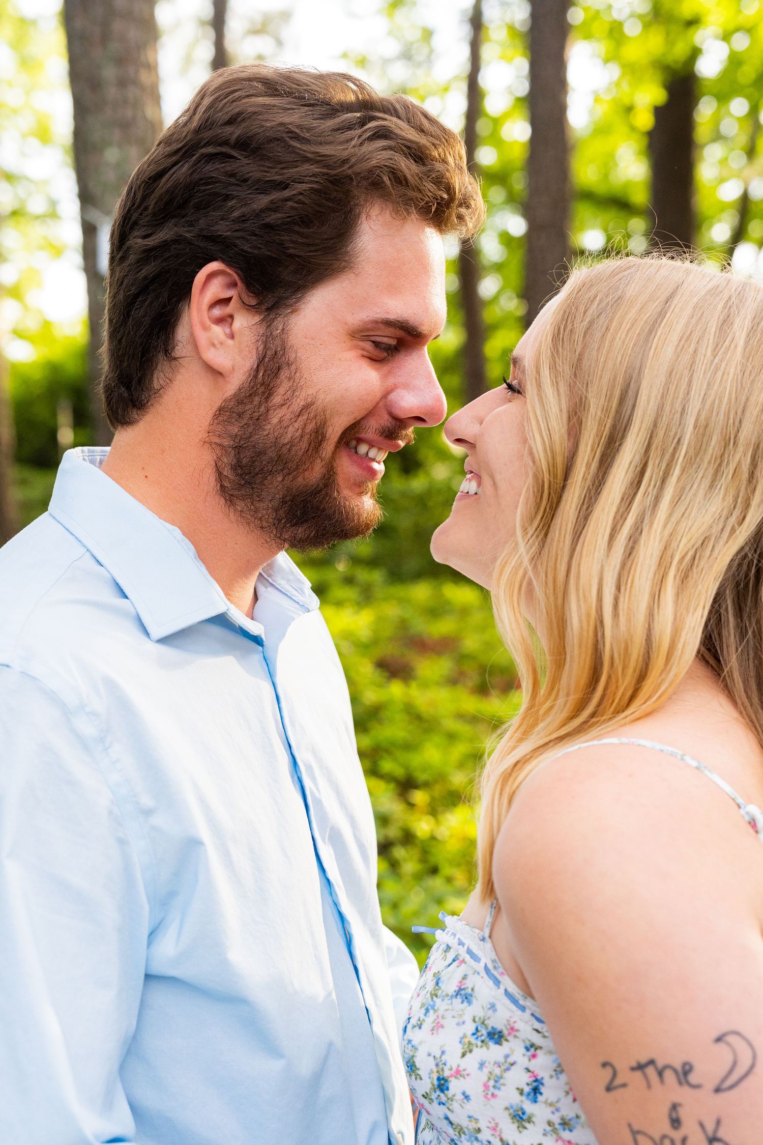 A couple shares a joyful intimate moment while smiling at each other outdoors in a sunlit wooded setting.