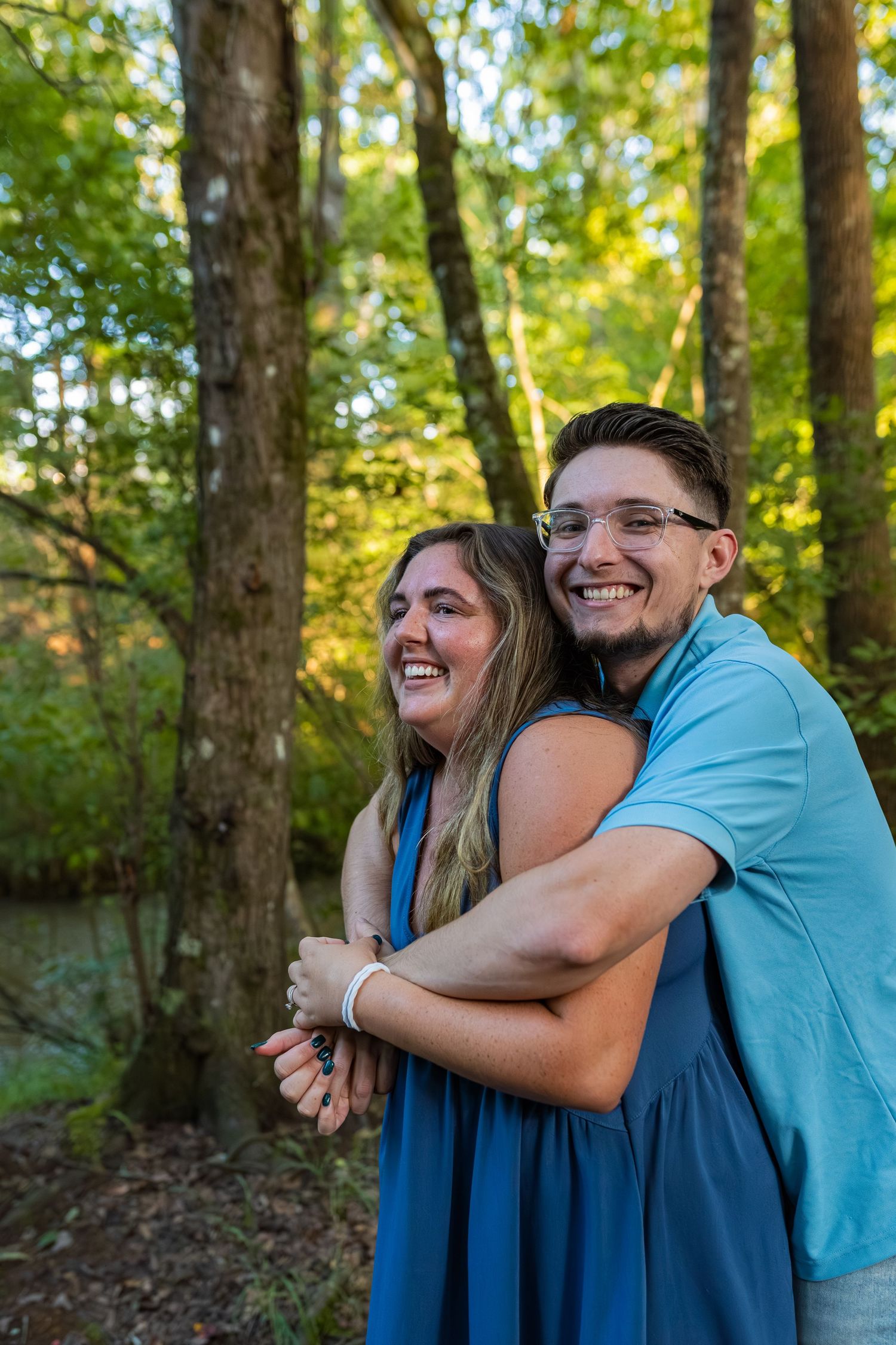 A couple shares a joyful embrace while standing in a sunlit forest setting with green trees in the background.