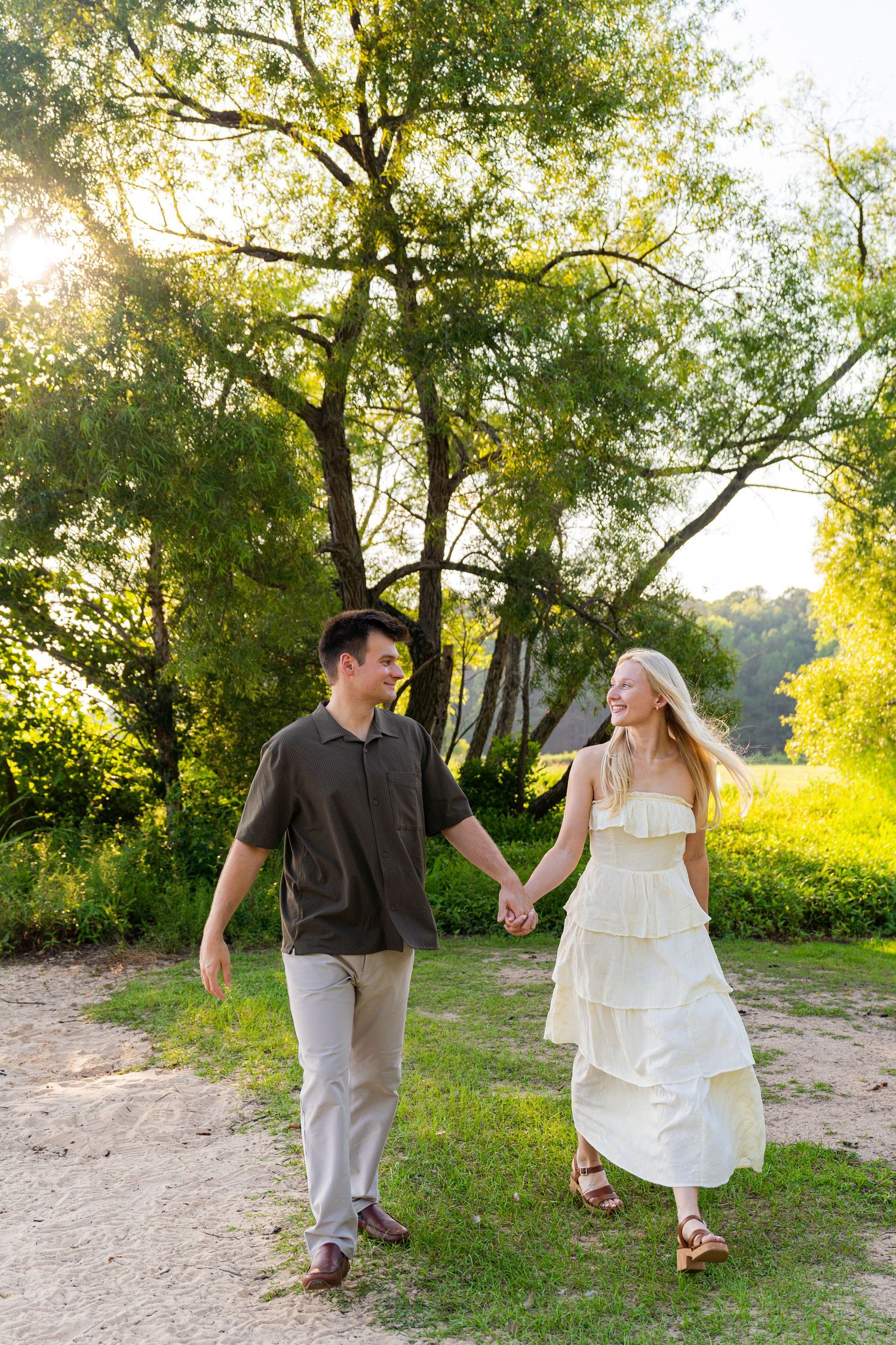 A couple holds hands and walks through a sunlit green park, smiling at each other, dressed in casual summer outfits.