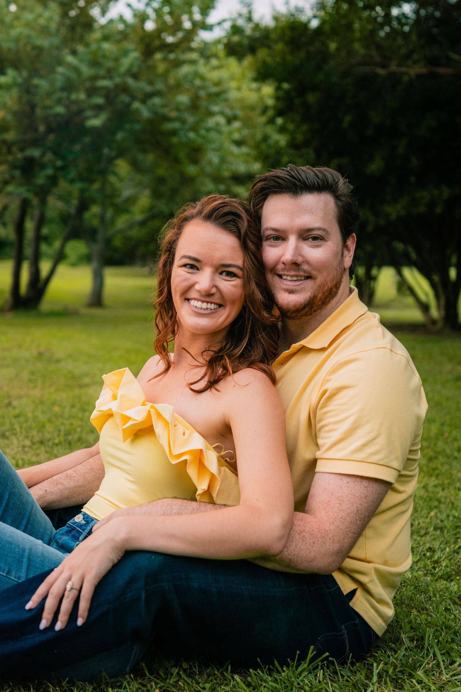A couple wearing yellow and jeans sit together on grass in a sunny park setting.