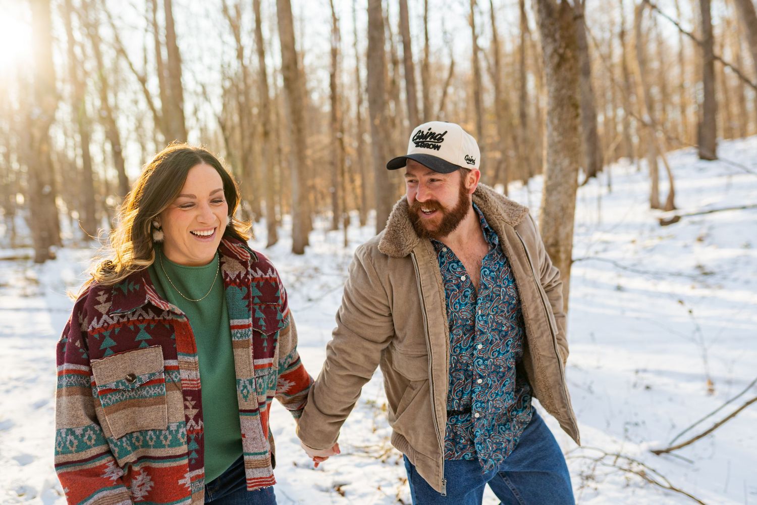 A couple in winter sweaters walks through a snowy forest path while holding hands and smiling together.