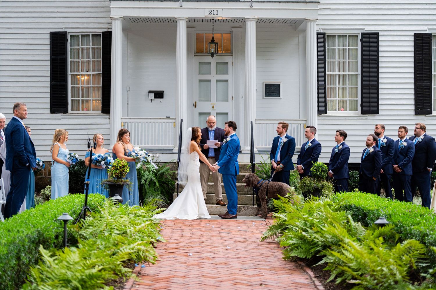 A wedding party gathers outside a white colonial-style building with brick pathways and lush green ferns.