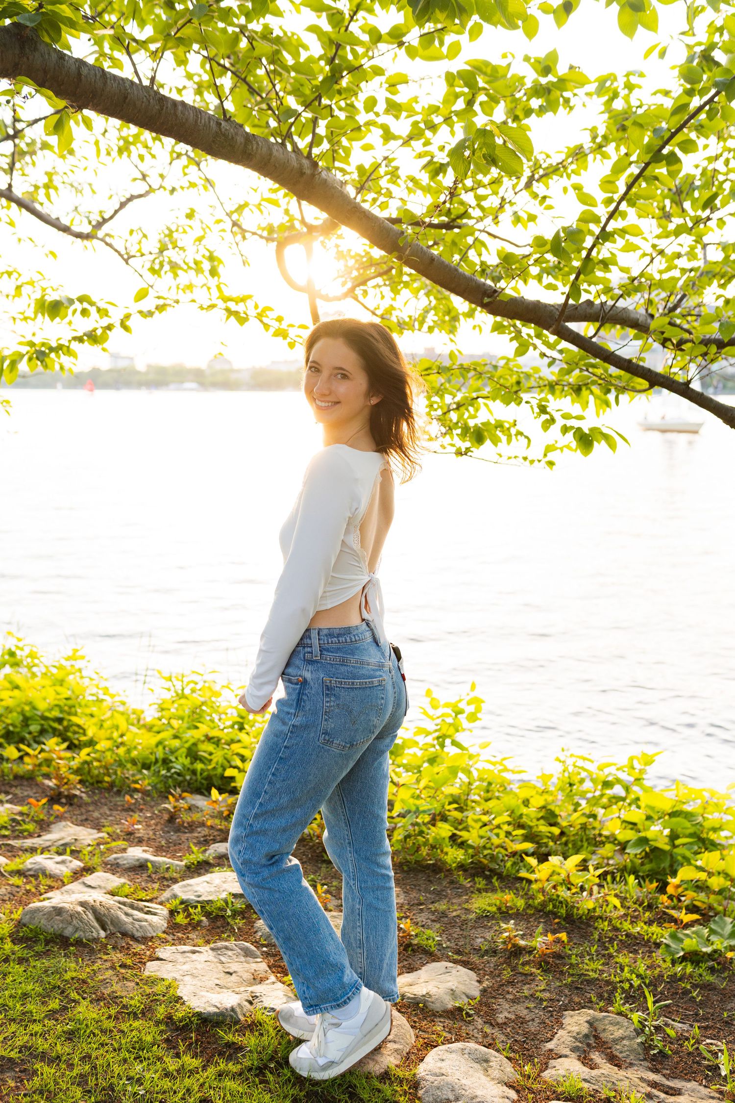 Person in casual outfit posing near water at sunset with green foliage overhead and rocks along shoreline.