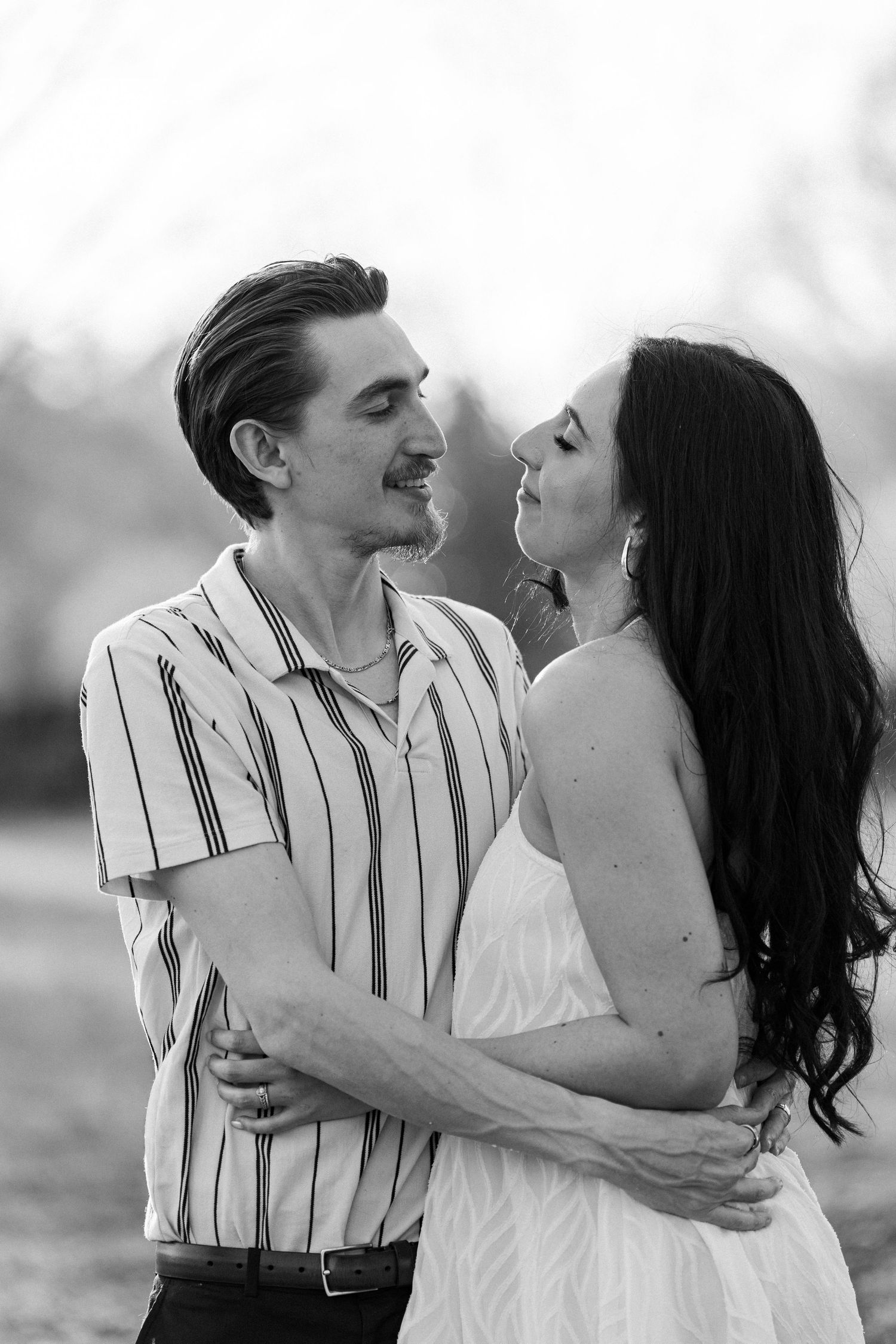 Black and white photo of a couple embracing and gazing at each other outdoors, man in striped shirt, woman in white dress.