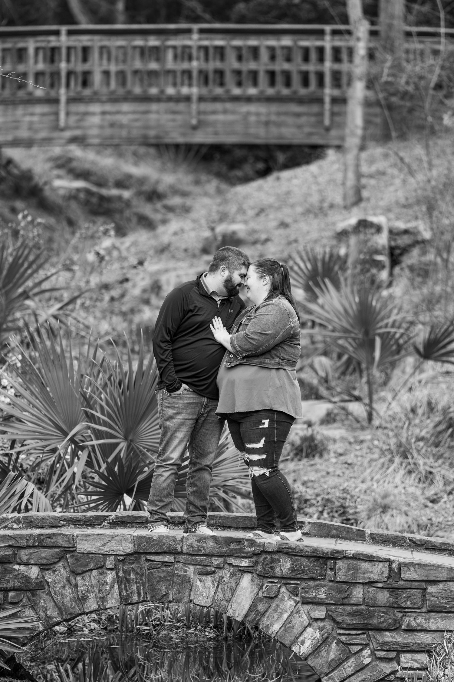 A romantic black and white photo of a couple sharing a moment on a stone bridge with palm plants in the background.