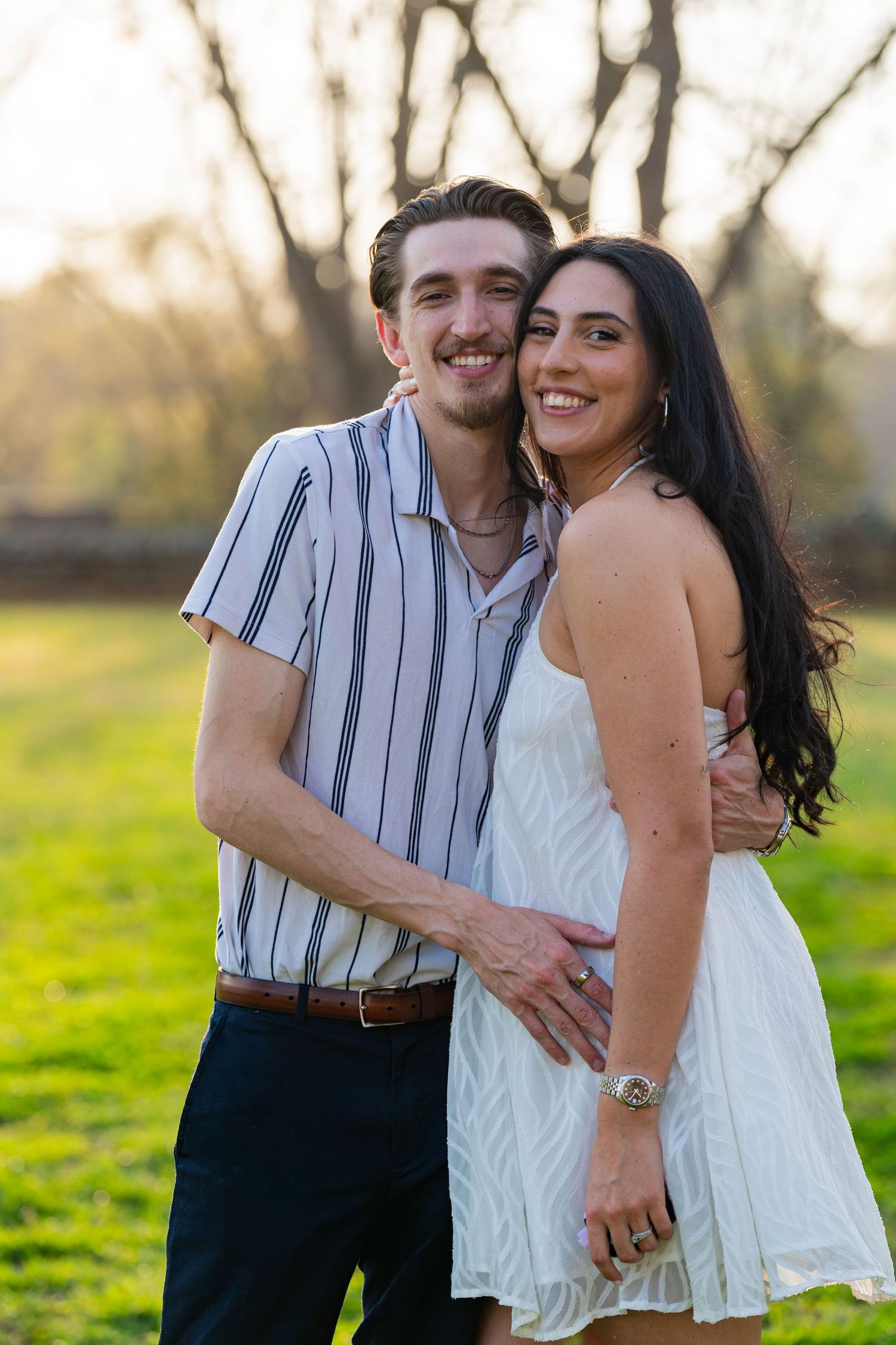 A smiling couple embraces outdoors on green grass with golden sunlight, man in striped shirt, woman in white dress.