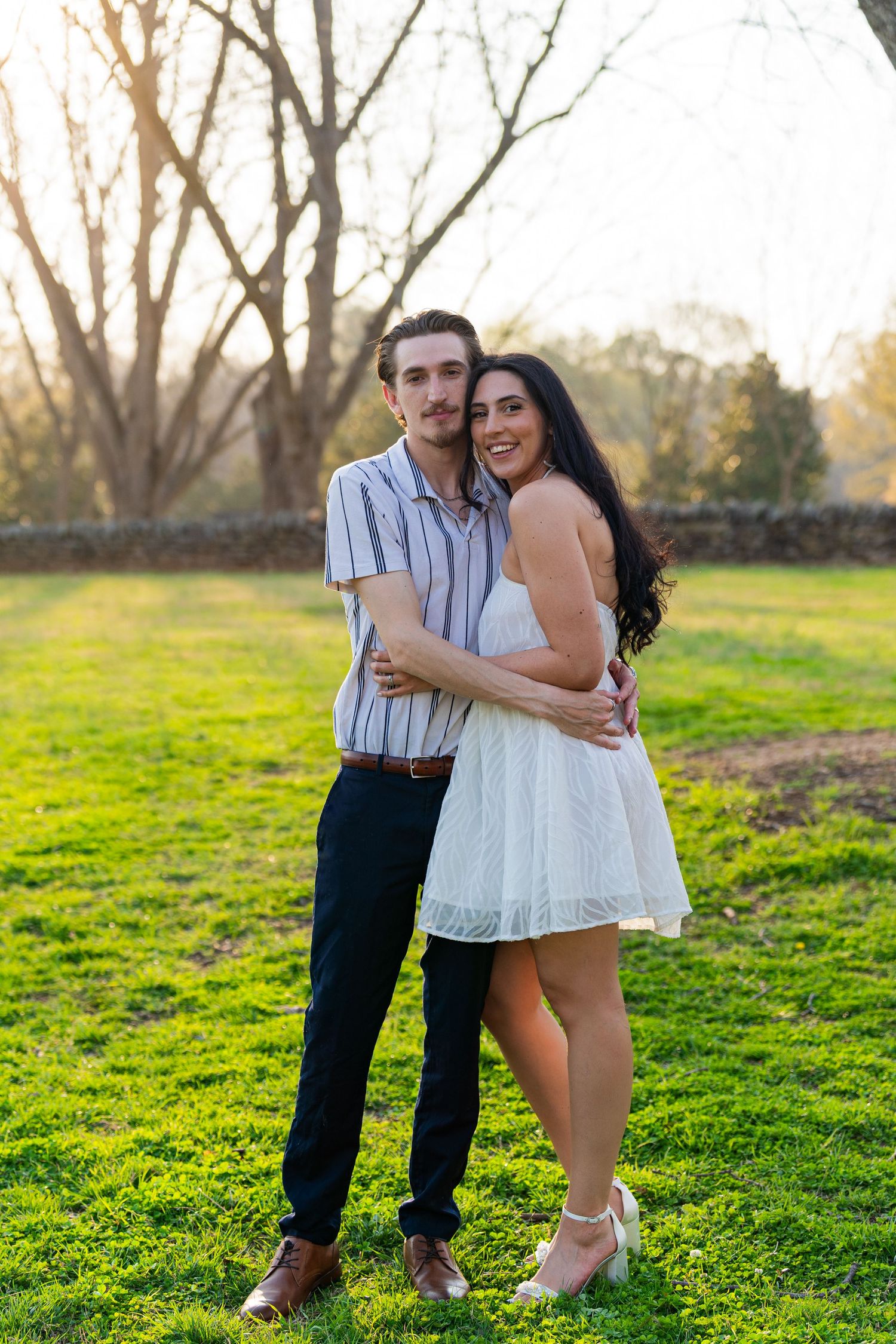 Couple embracing outdoors on green grass at golden hour, woman in white dress and man in striped shirt smiling.