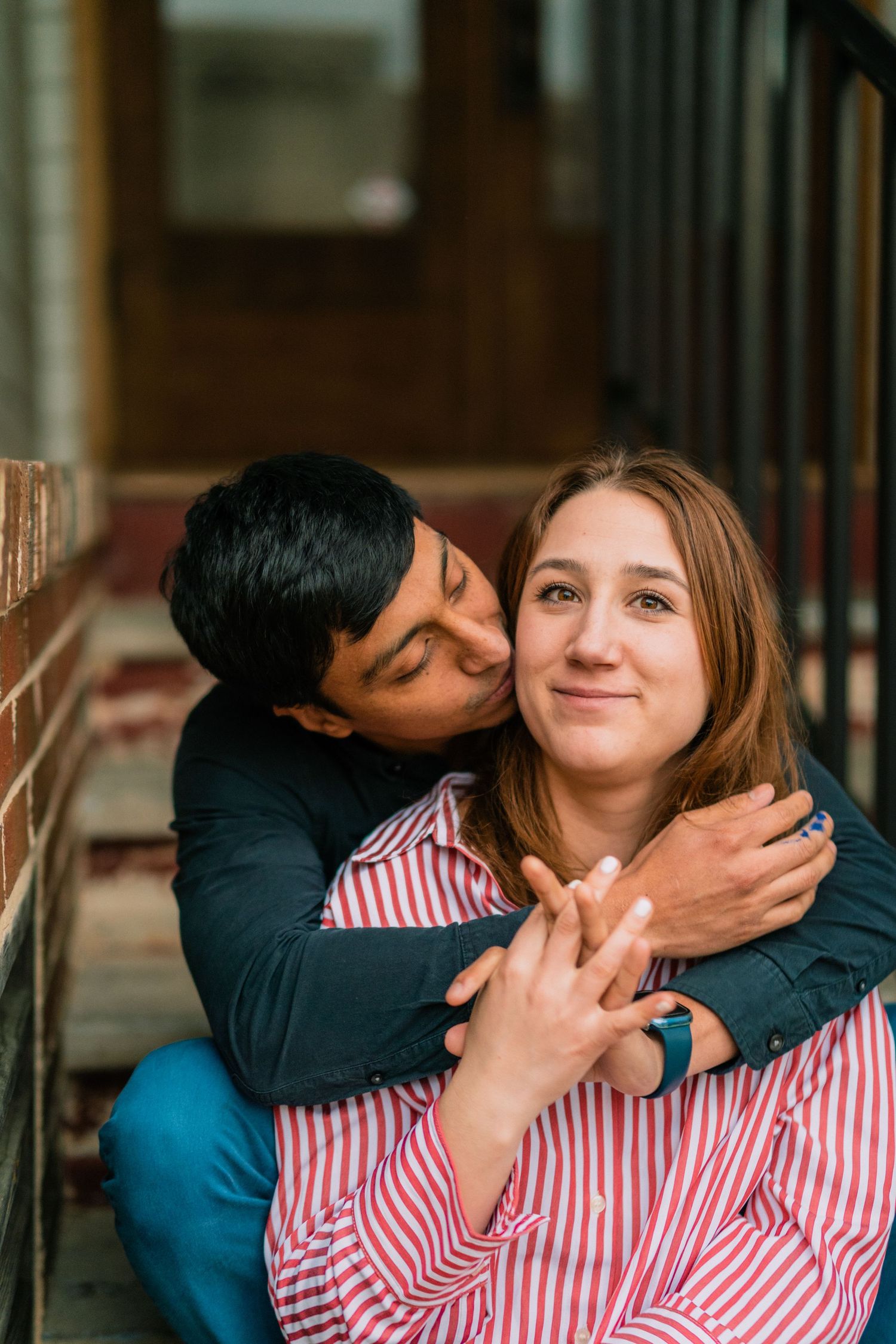 A couple sharing an affectionate moment on stairs, with one wearing a striped red and white shirt.