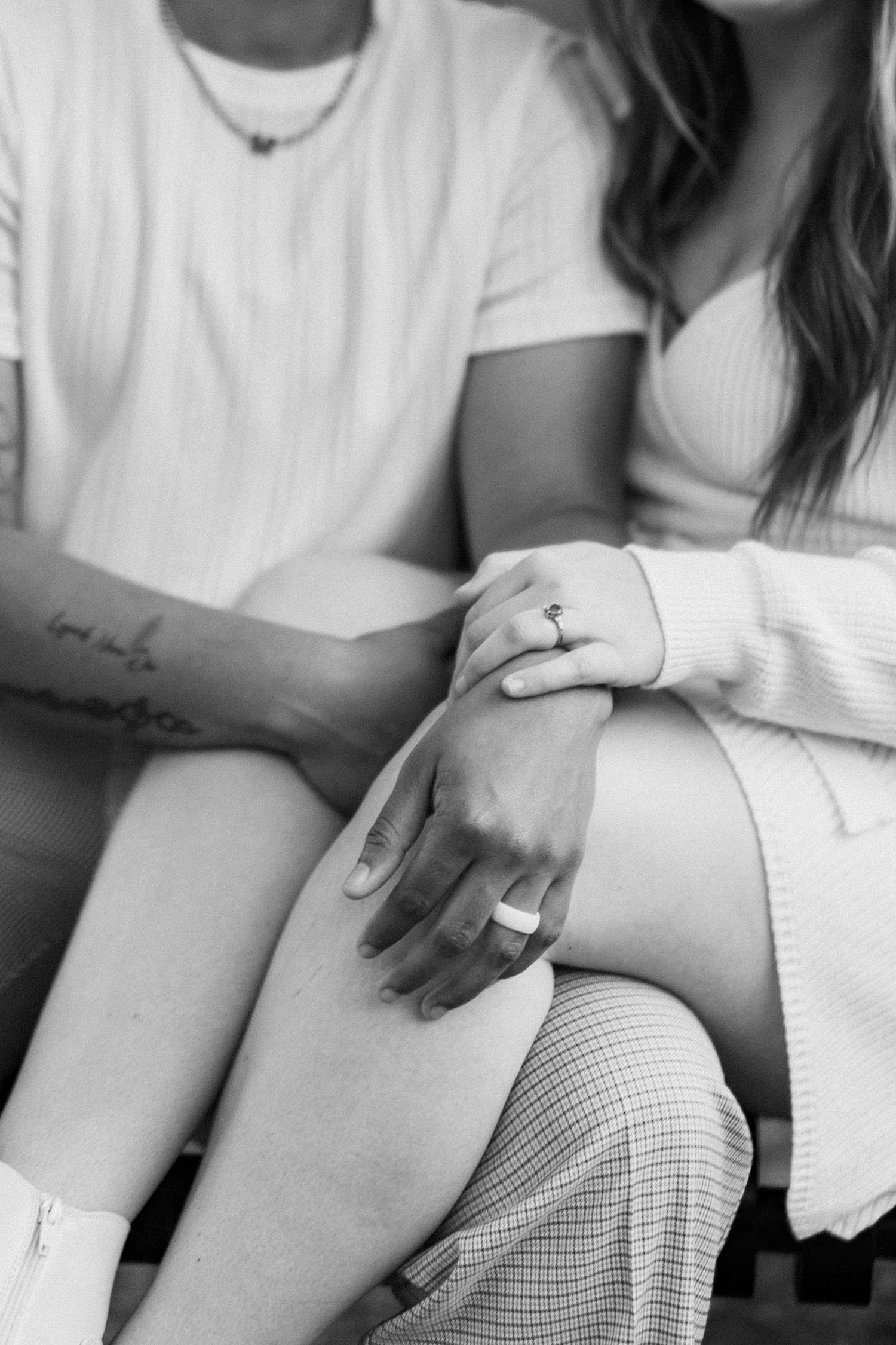 A close-up black and white photo showing two people's hands with rings resting on their legs.