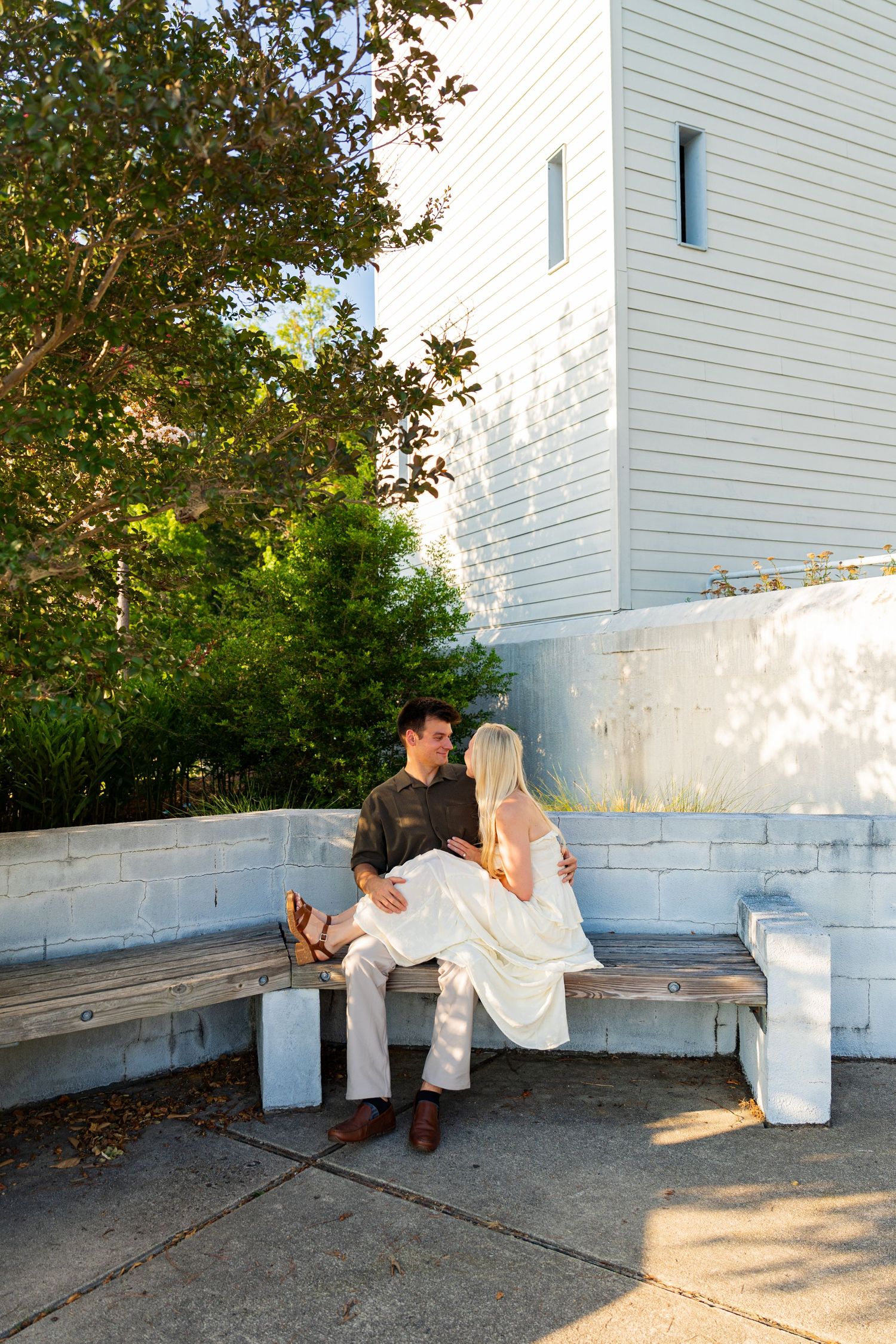 Couple sitting together on an outdoor bench near a white building, surrounded by lush green trees in warm sunlight.