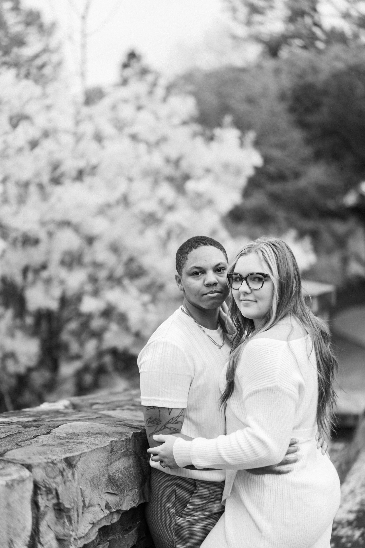 A couple in white clothing embracing while posing against a stone wall outdoors.