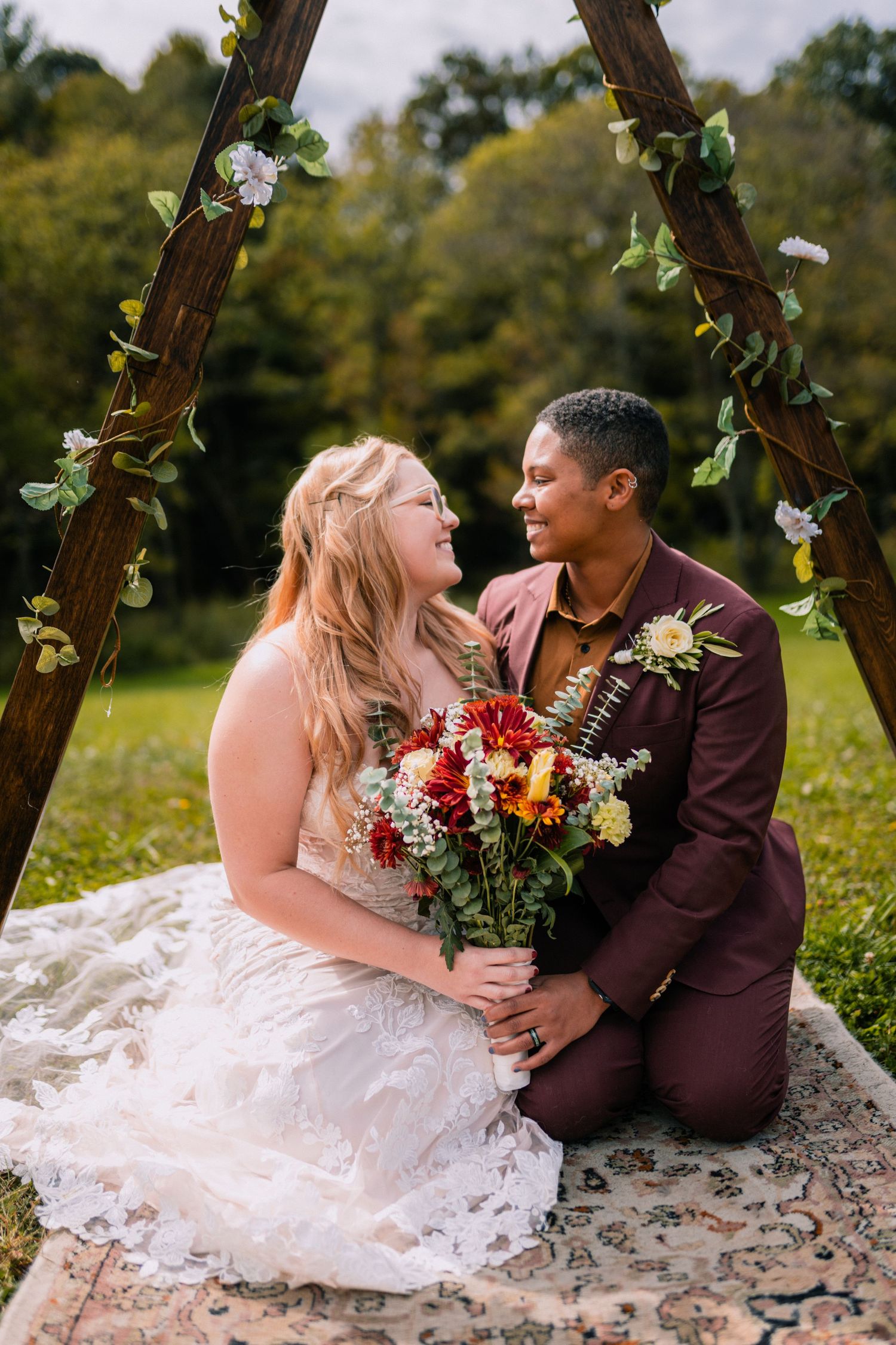 Two newlyweds share a romantic moment on a blanket under a floral archway in an outdoor garden setting.