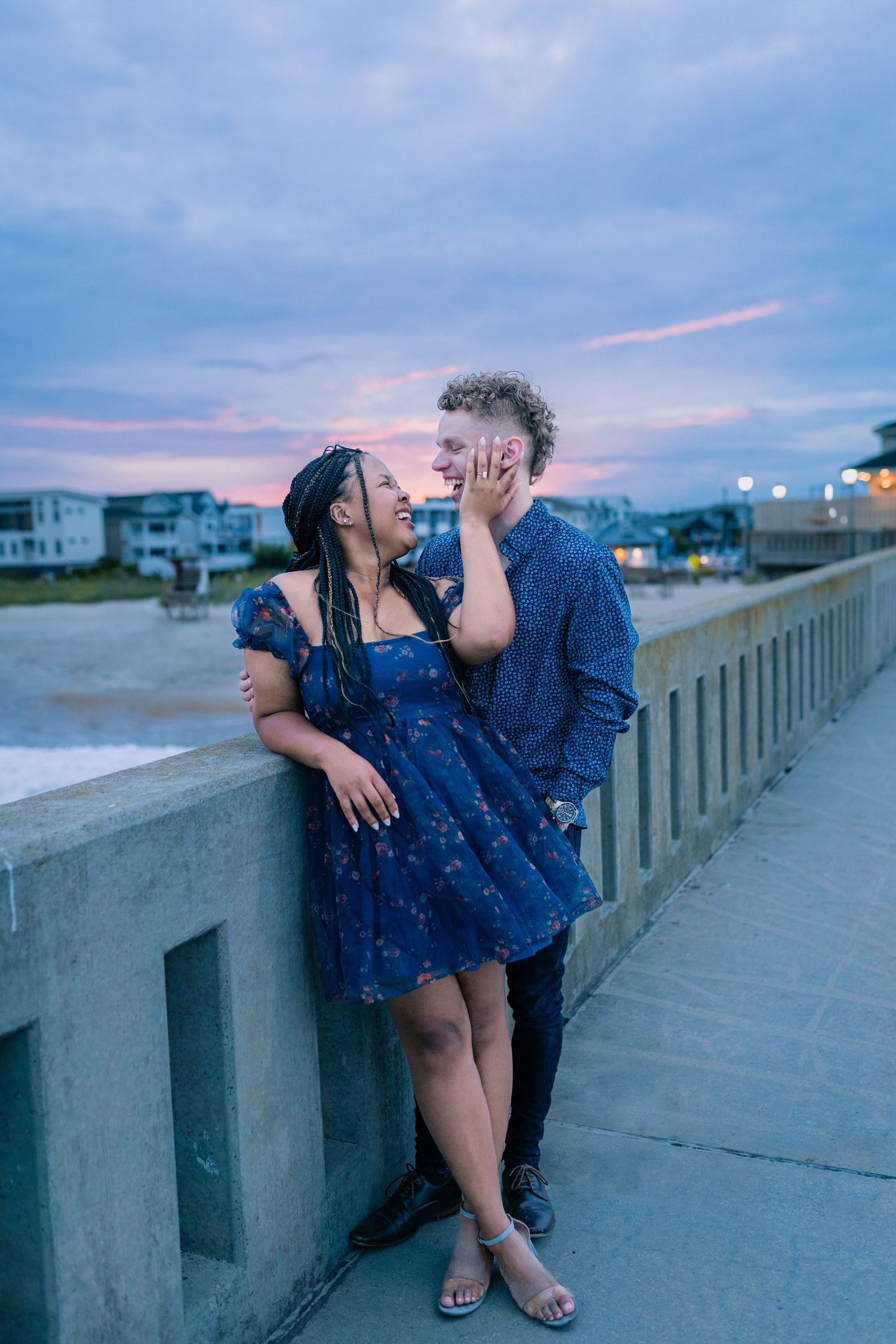 A romantic couple in matching navy blue outfits share a tender moment at sunset along a coastal boardwalk.