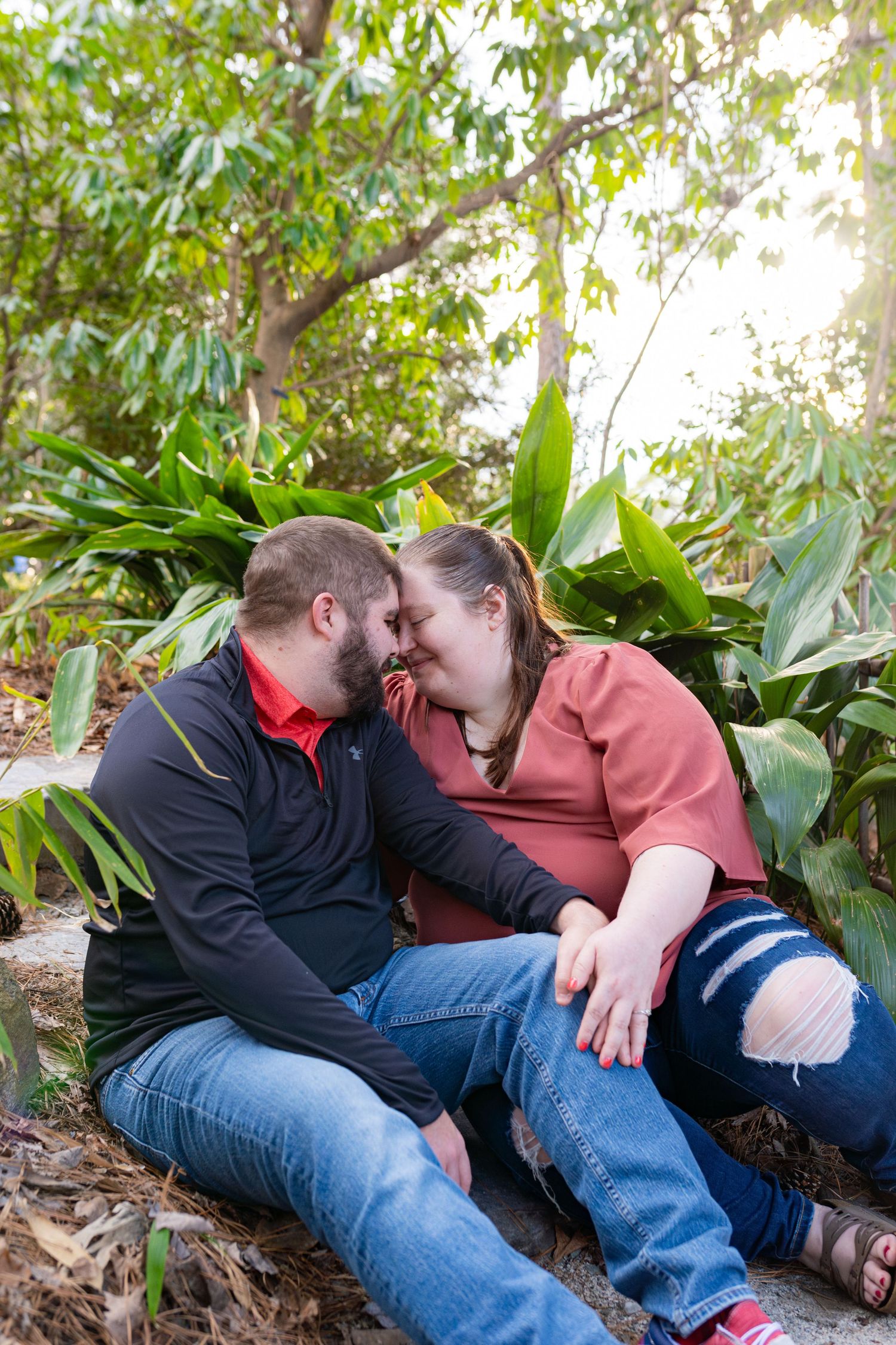 A couple shares an intimate moment while sitting together on fallen leaves in a garden setting.