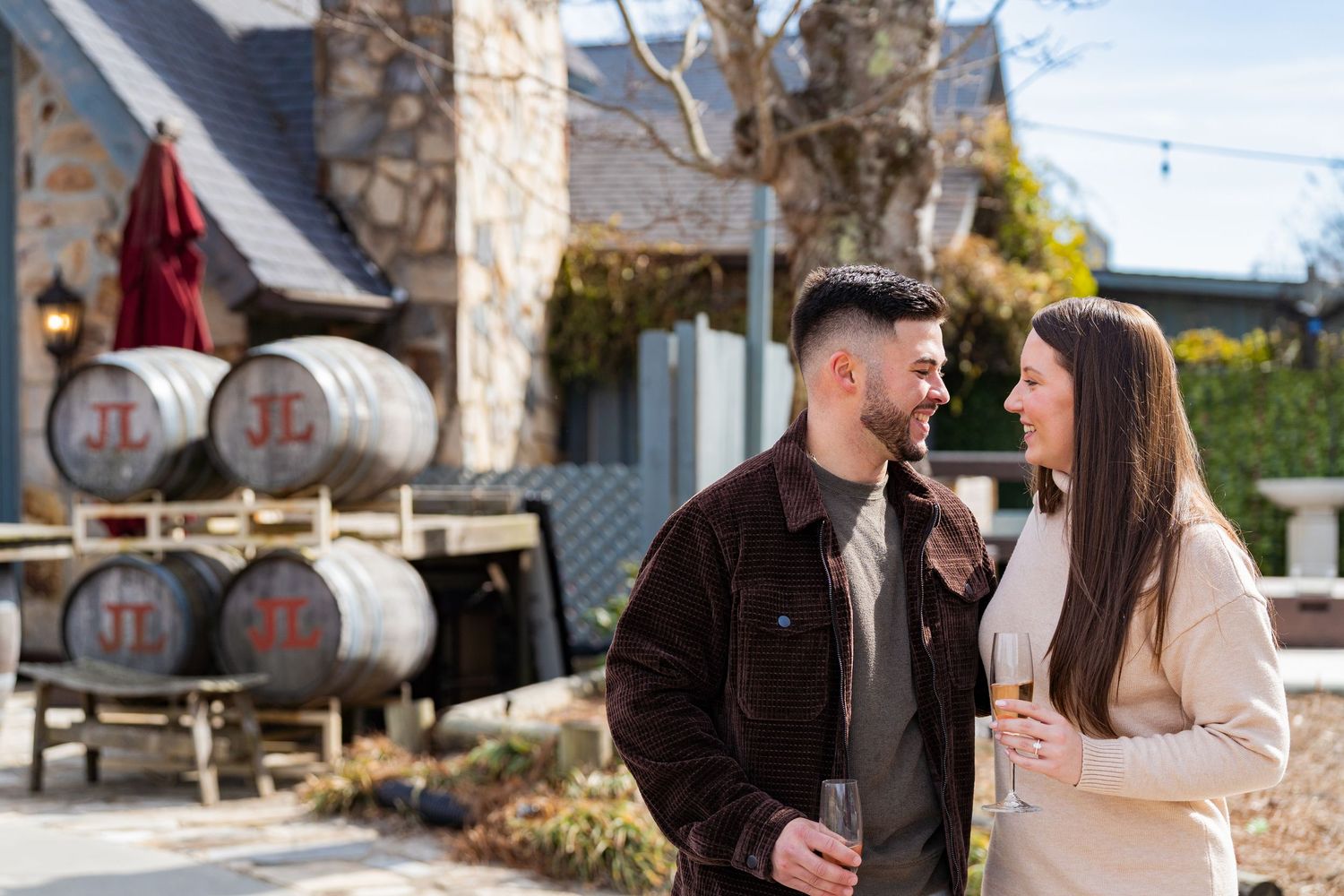 A couple enjoys wine together at a rustic winery with wine barrels in the background on a sunny day.