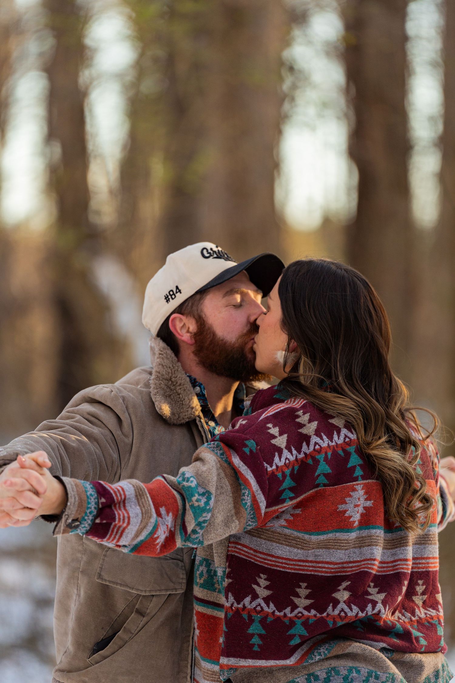 A couple wearing matching southwestern patterned sweaters share a romantic kiss in a snowy winter forest setting.