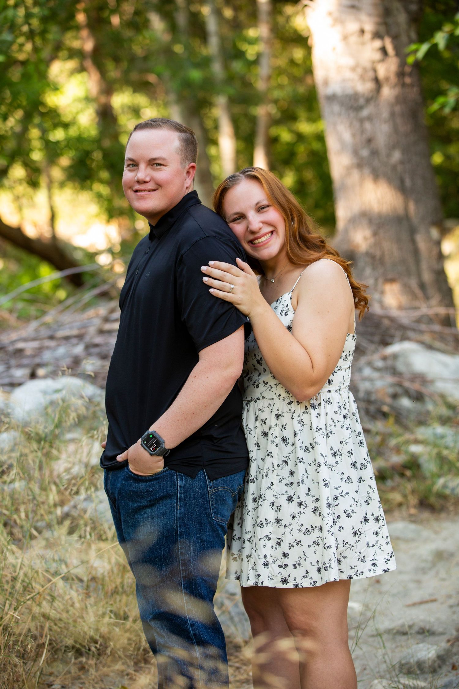 A happy couple embraces and laughs together during an outdoor photoshoot among trees and rocks.