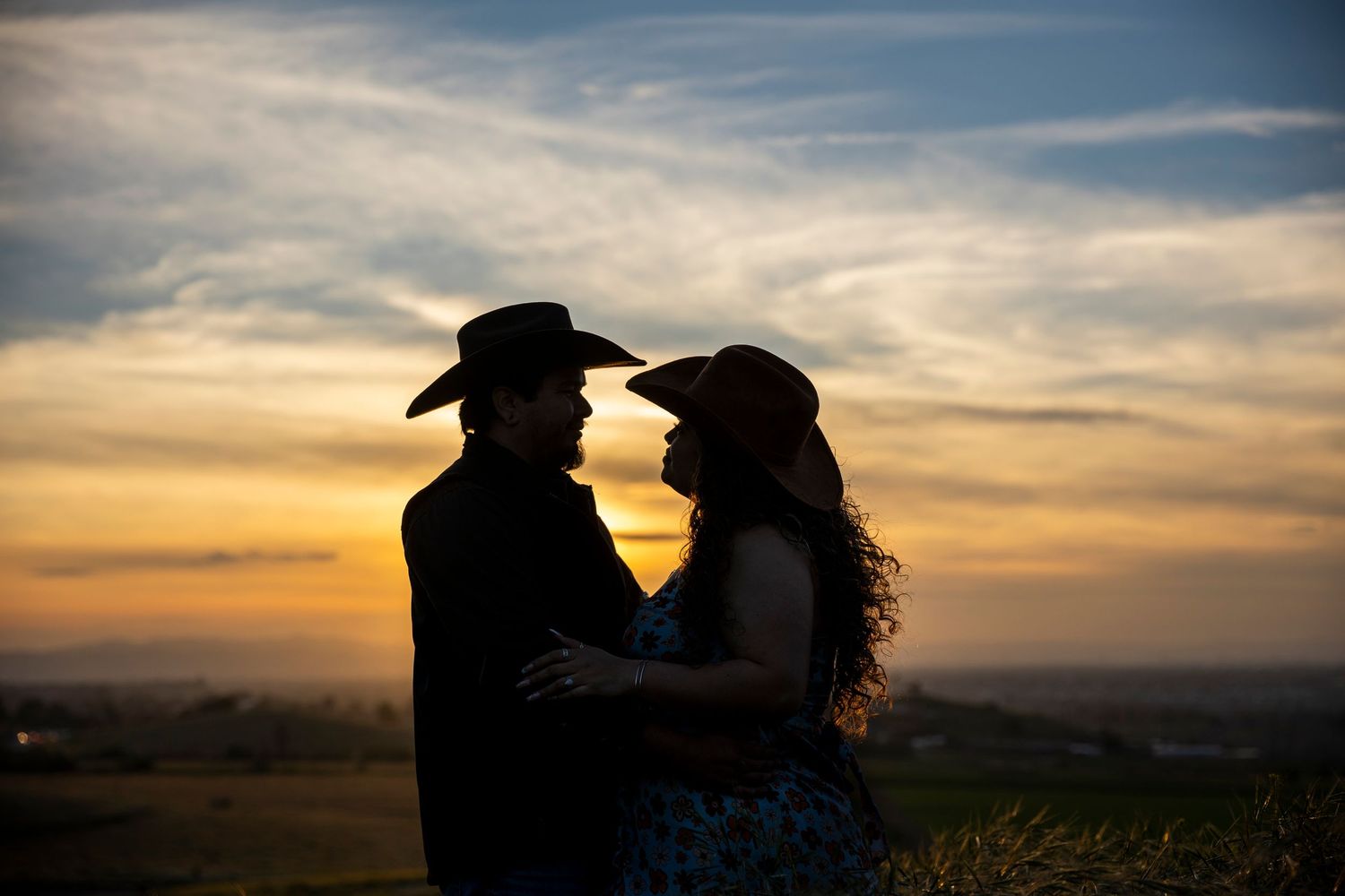 A romantic couple in silhouette stands close together against a glowing sunset sky over rolling hills.