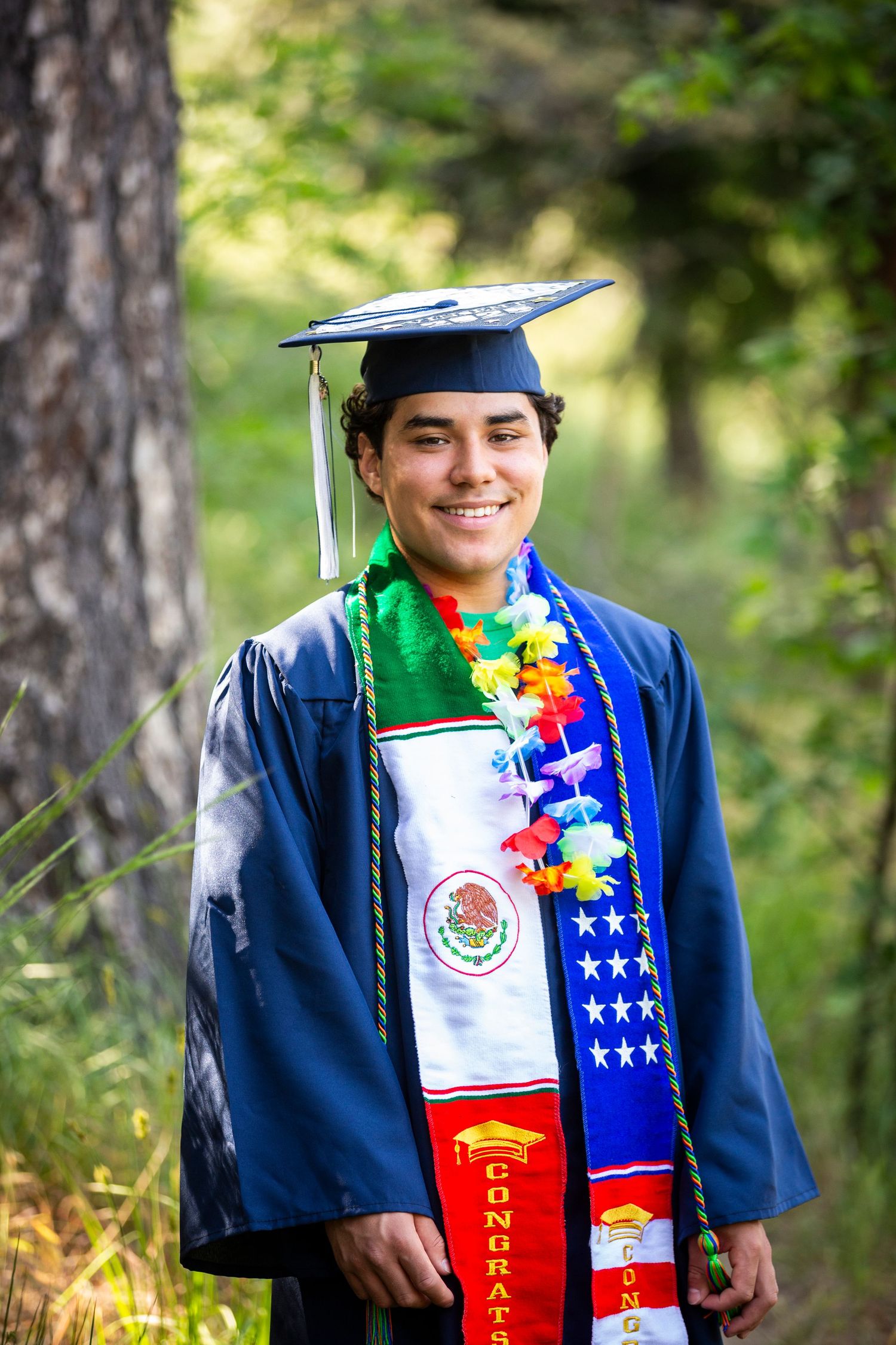 Graduate wearing blue academic regalia and colorful stoles poses outdoors in nature for graduation photos.
