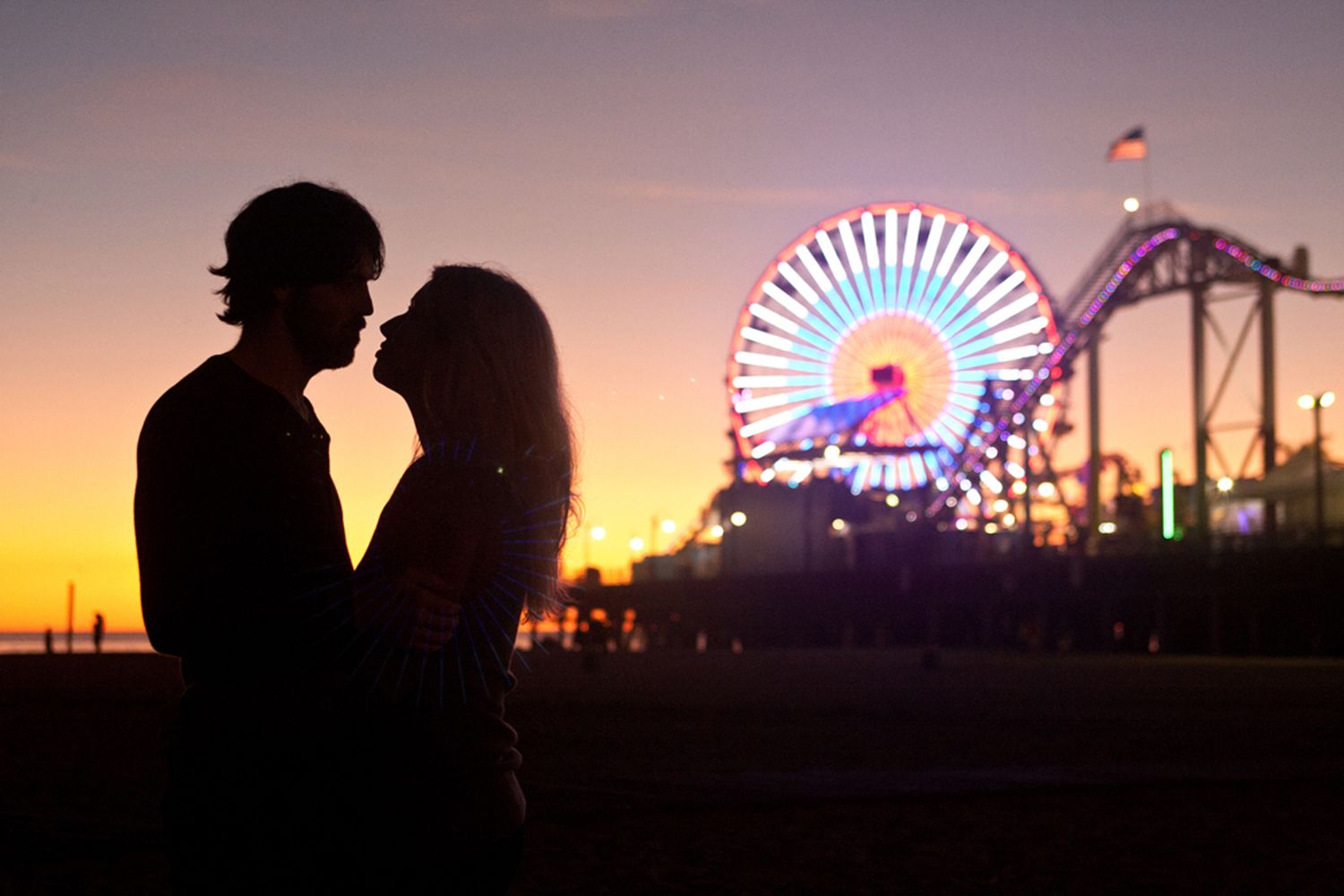 Silhouetted couple against sunset sky with illuminated Ferris wheel and amusement park in background.