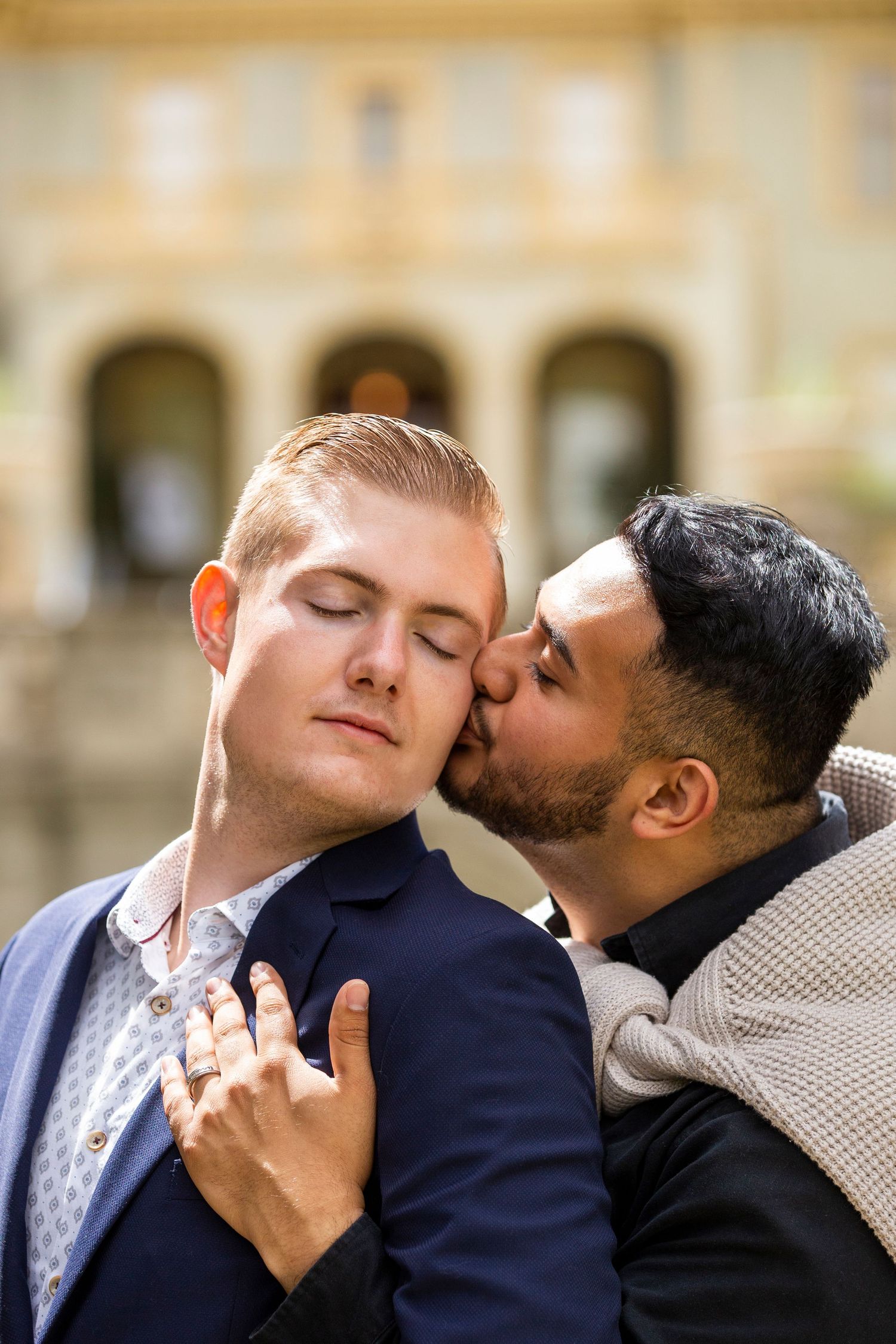Two people share an intimate moment during a romantic photoshoot in front of a building with elegant architecture.