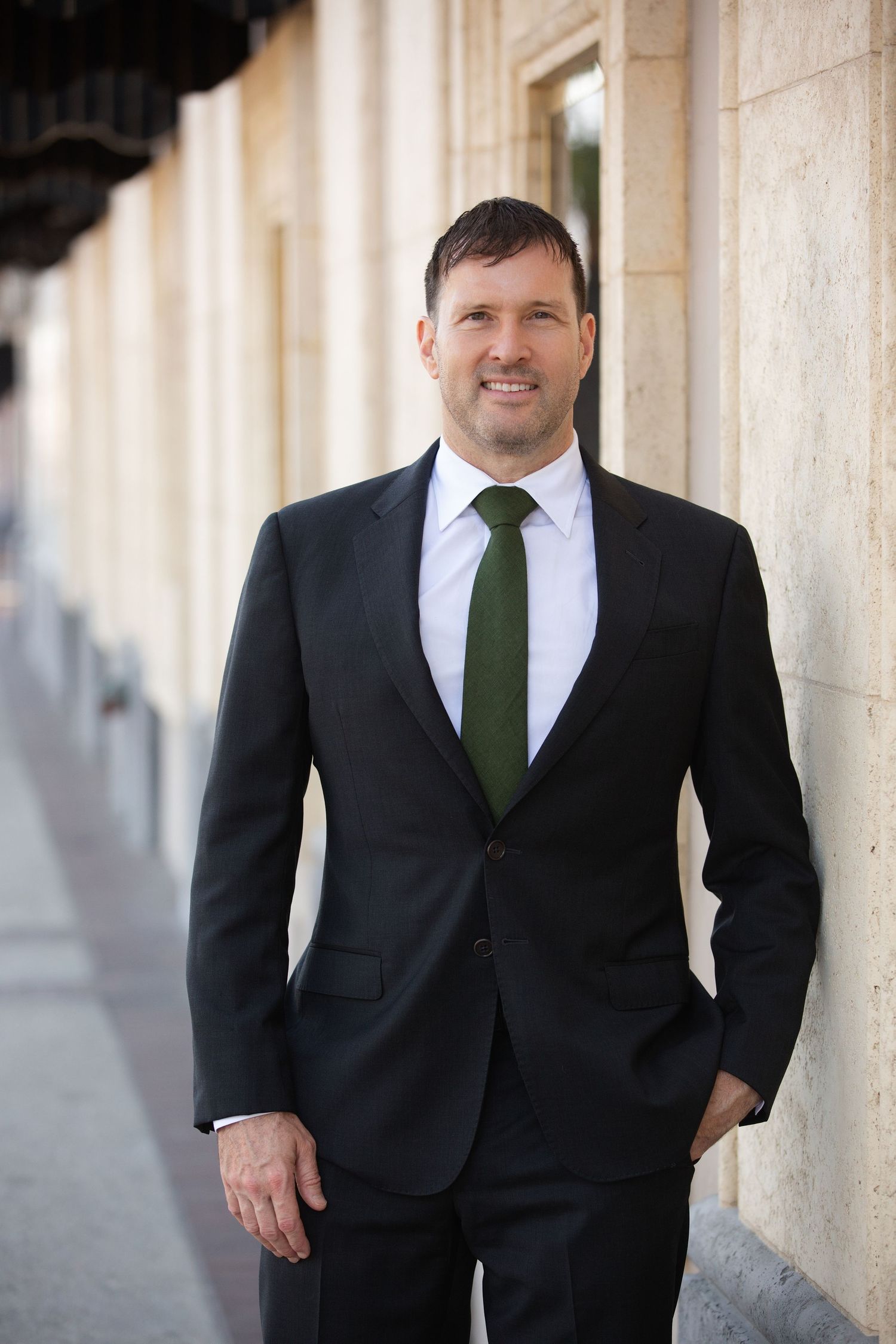 Professional wearing a dark suit with green tie stands confidently against a light stone building exterior.