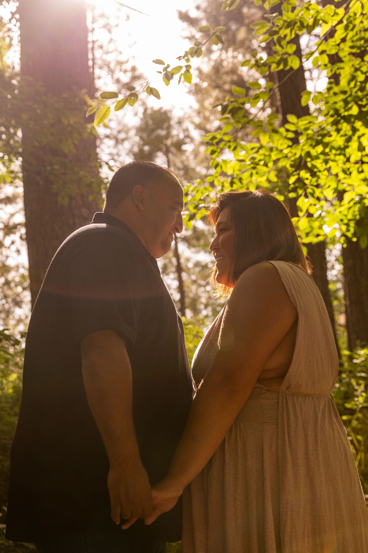 A couple stands together in sunlit woods during a romantic outdoor moment, holding hands and smiling at each other.