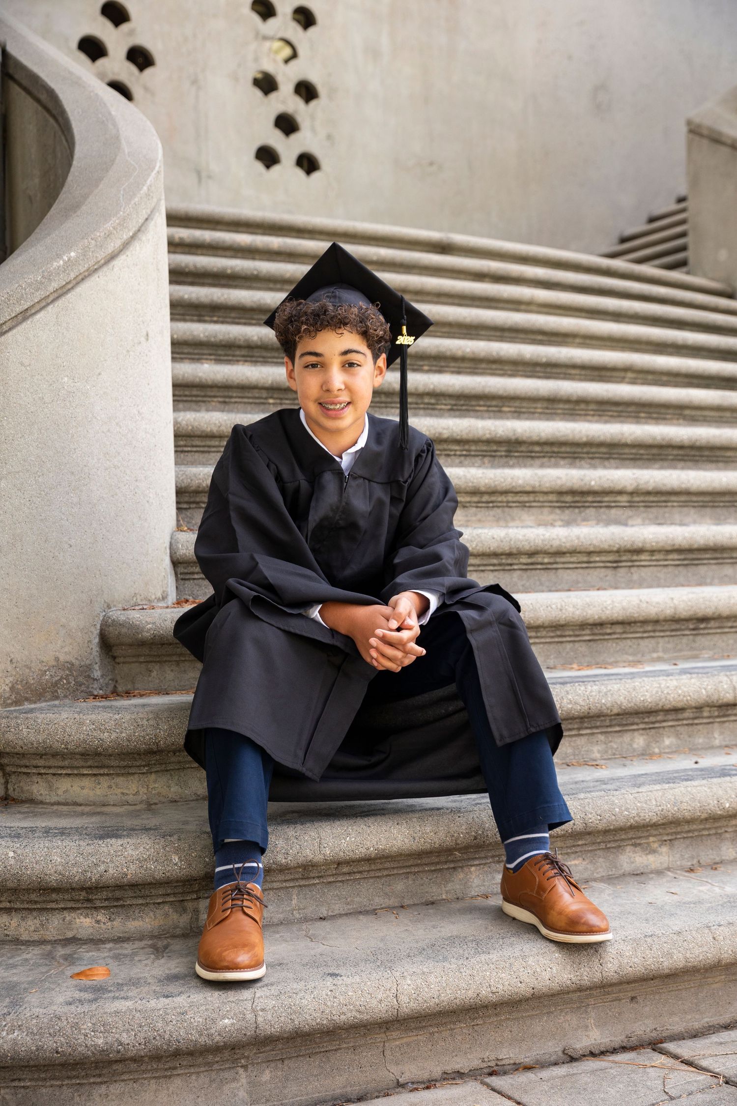 Graduate in black cap and gown sits casually on stone steps while using phone.