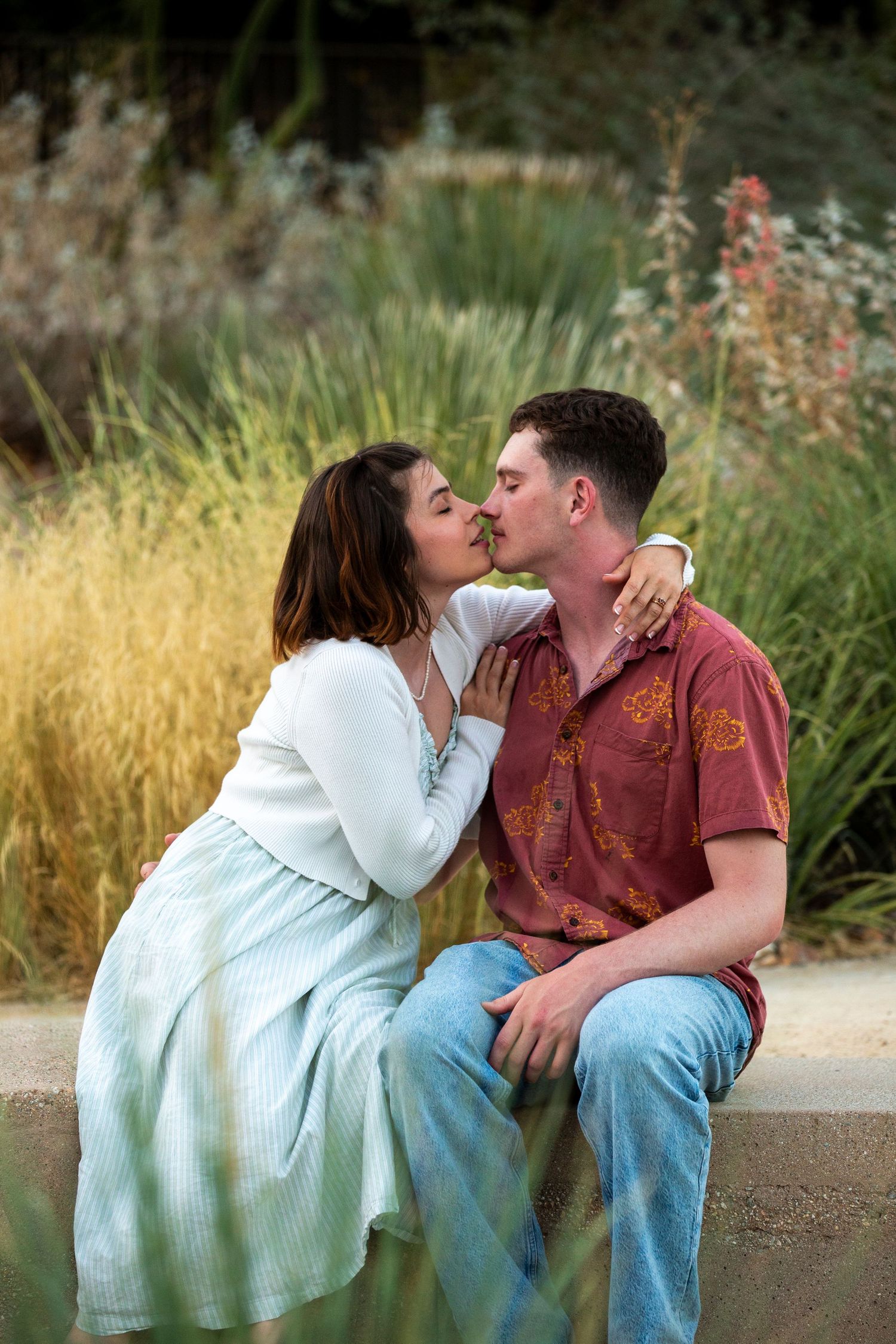 A romantic couple sharing a tender kiss while sitting outdoors among tall grasses during golden hour.