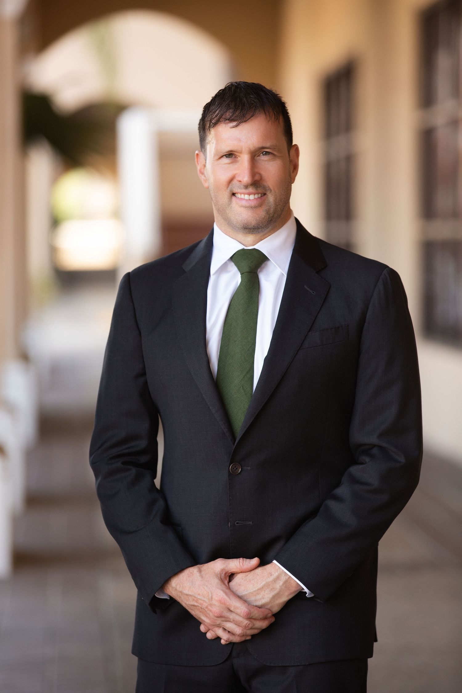 Professional wearing a black suit and dark green tie stands in a covered walkway with arched architecture.