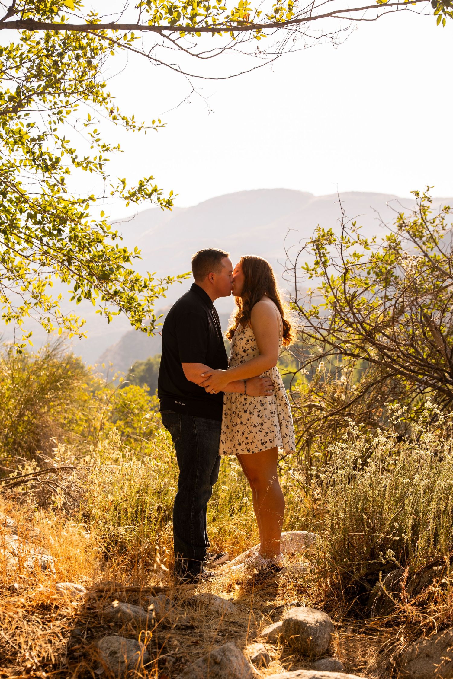 A couple shares a romantic kiss on a hillside trail surrounded by golden sunlit foliage and mountain views in the background.