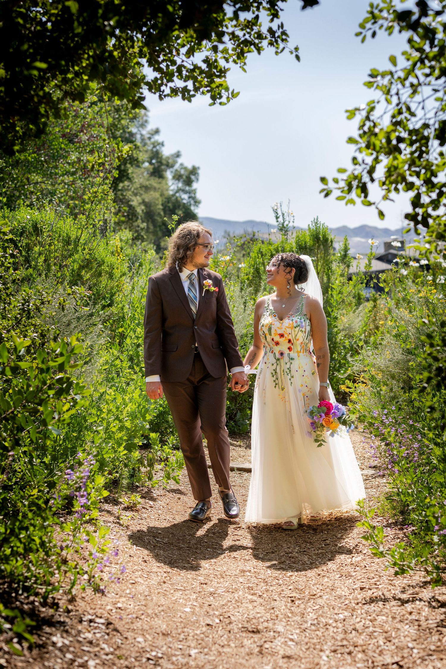 A newlywed couple walks together on a peaceful dirt path surrounded by green foliage in a sunlit outdoor setting.