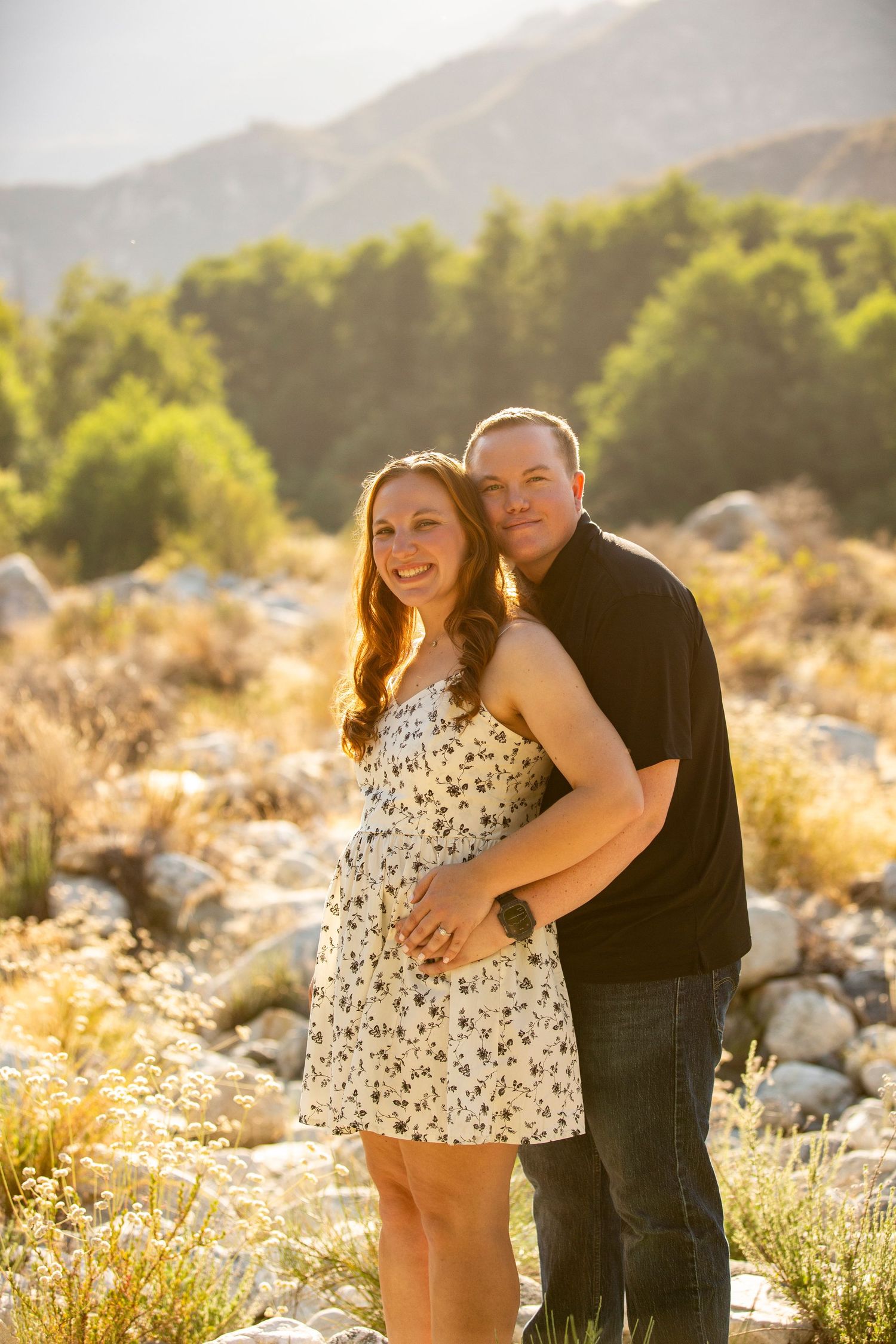 A couple poses together in a natural desert setting with mountains and sunlit foliage in the background.