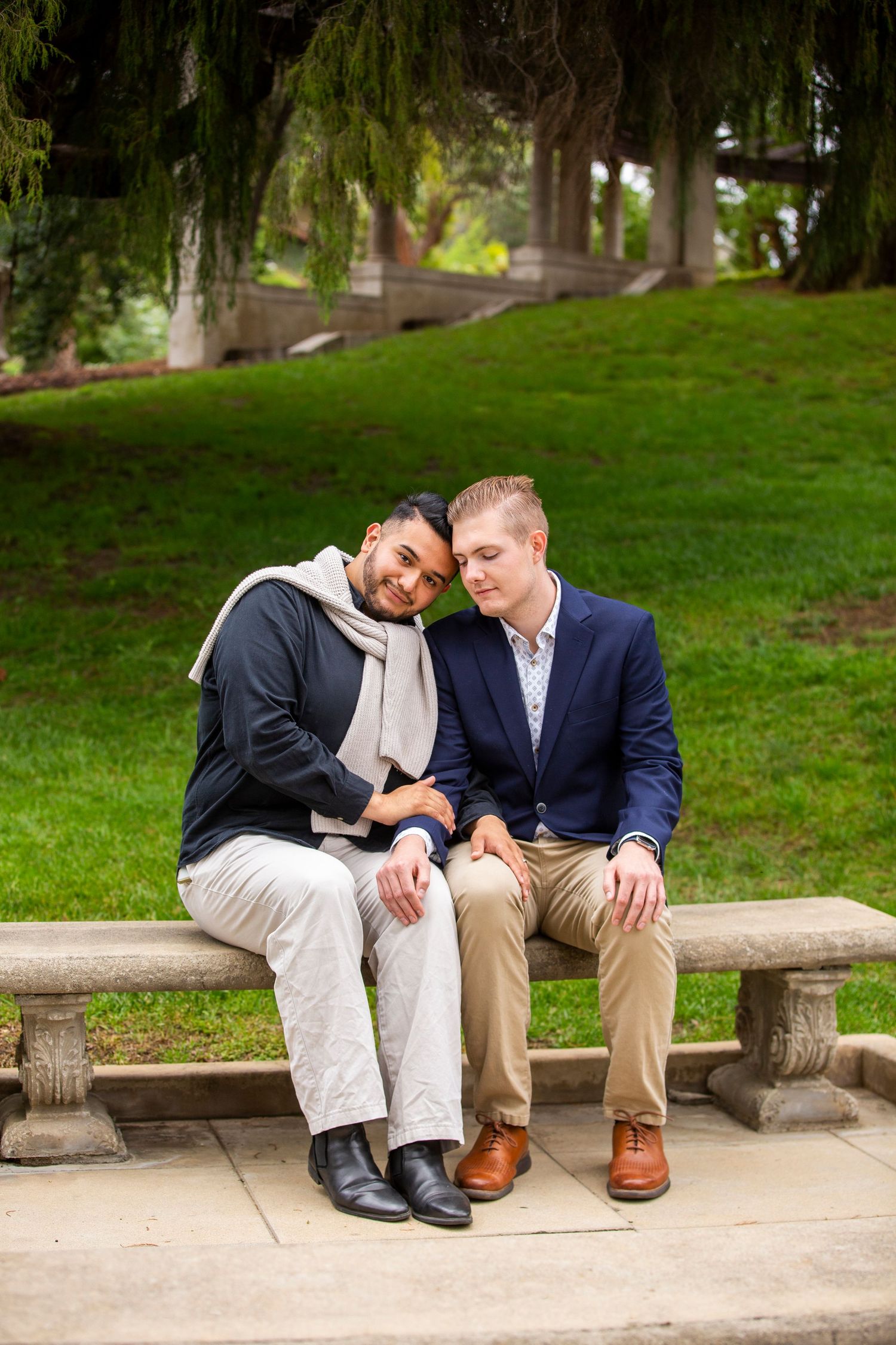 Two people share a sweet moment while sitting close together on a stone bench in a peaceful park setting.