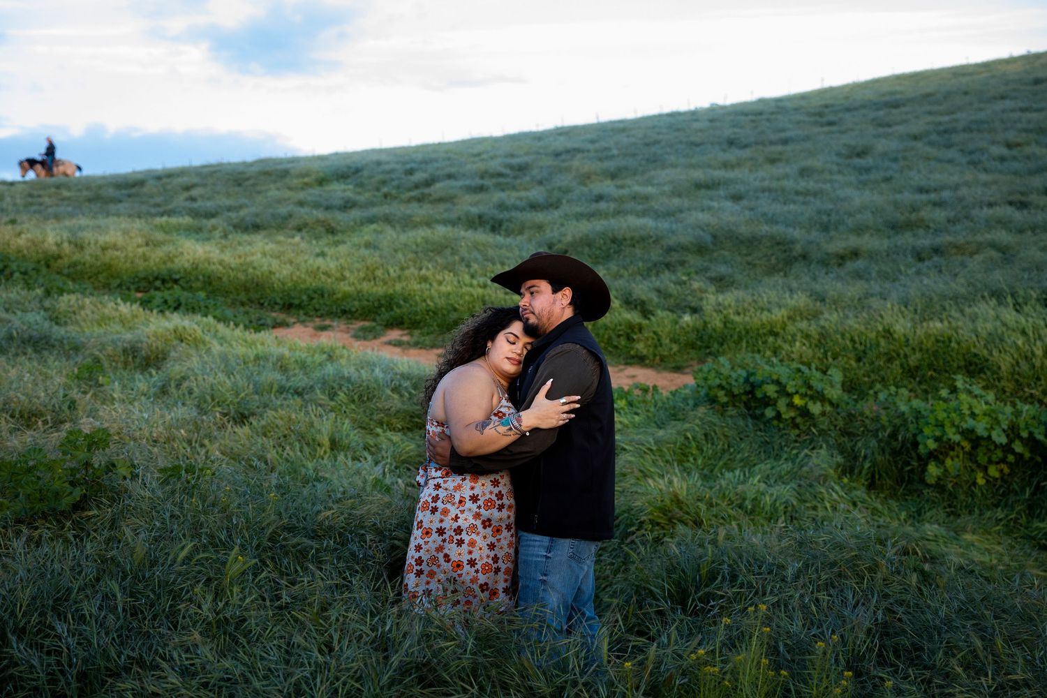 Two people embrace while standing in green rolling hills against a blue sky background.