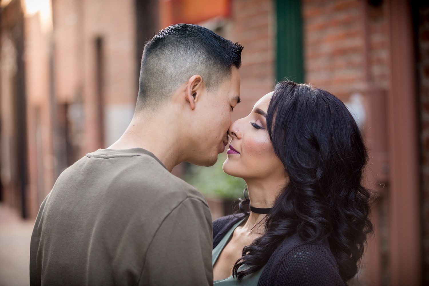 A couple wearing fall colors stands close together with textured brick walls in the background.