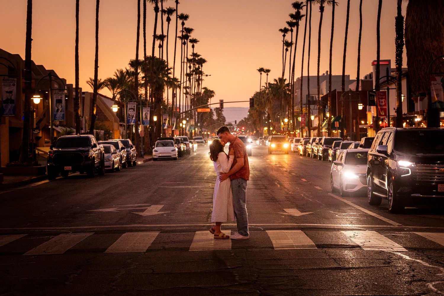 A couple embraces in the middle of a palm tree-lined street at sunset with car headlights glowing in California.