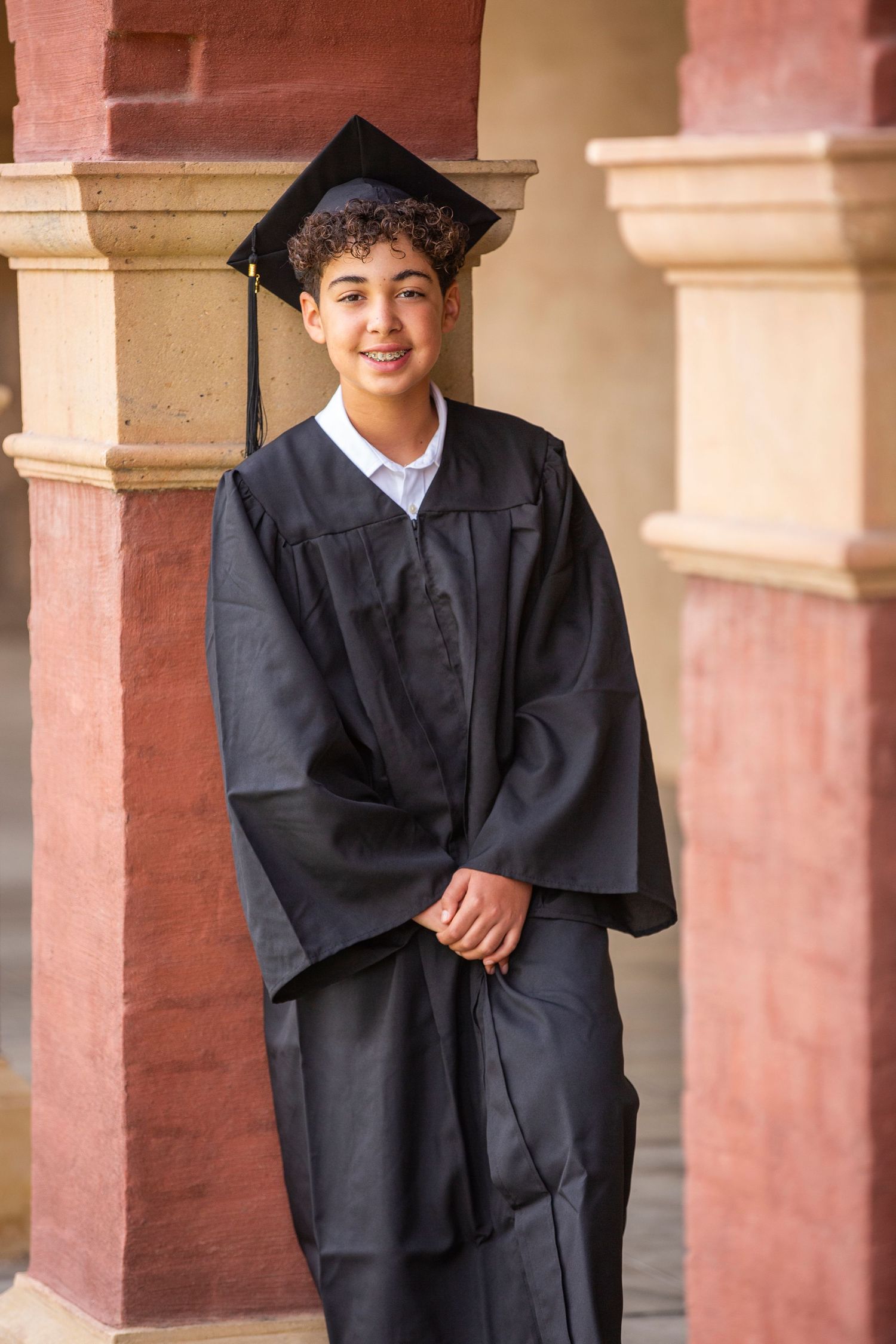 Graduate in black cap and gown standing and smiling between pink stone pillars during graduation photoshoot.