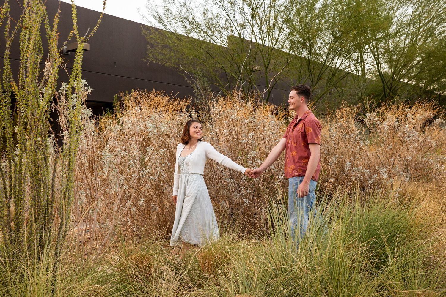 Couple playfully interacting in a rustic outdoor setting with tall grass and autumn foliage at sunset.