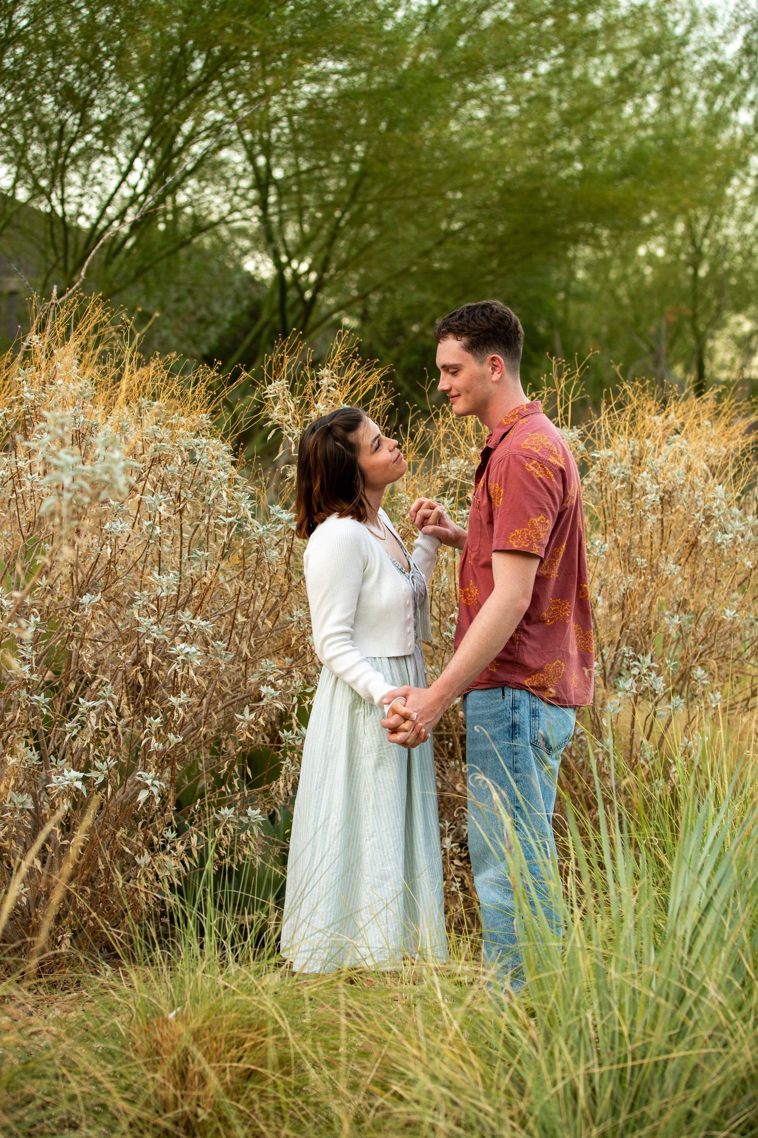 A couple shares a tender moment amid tall grass and golden wildflowers during an outdoor photoshoot at sunset.