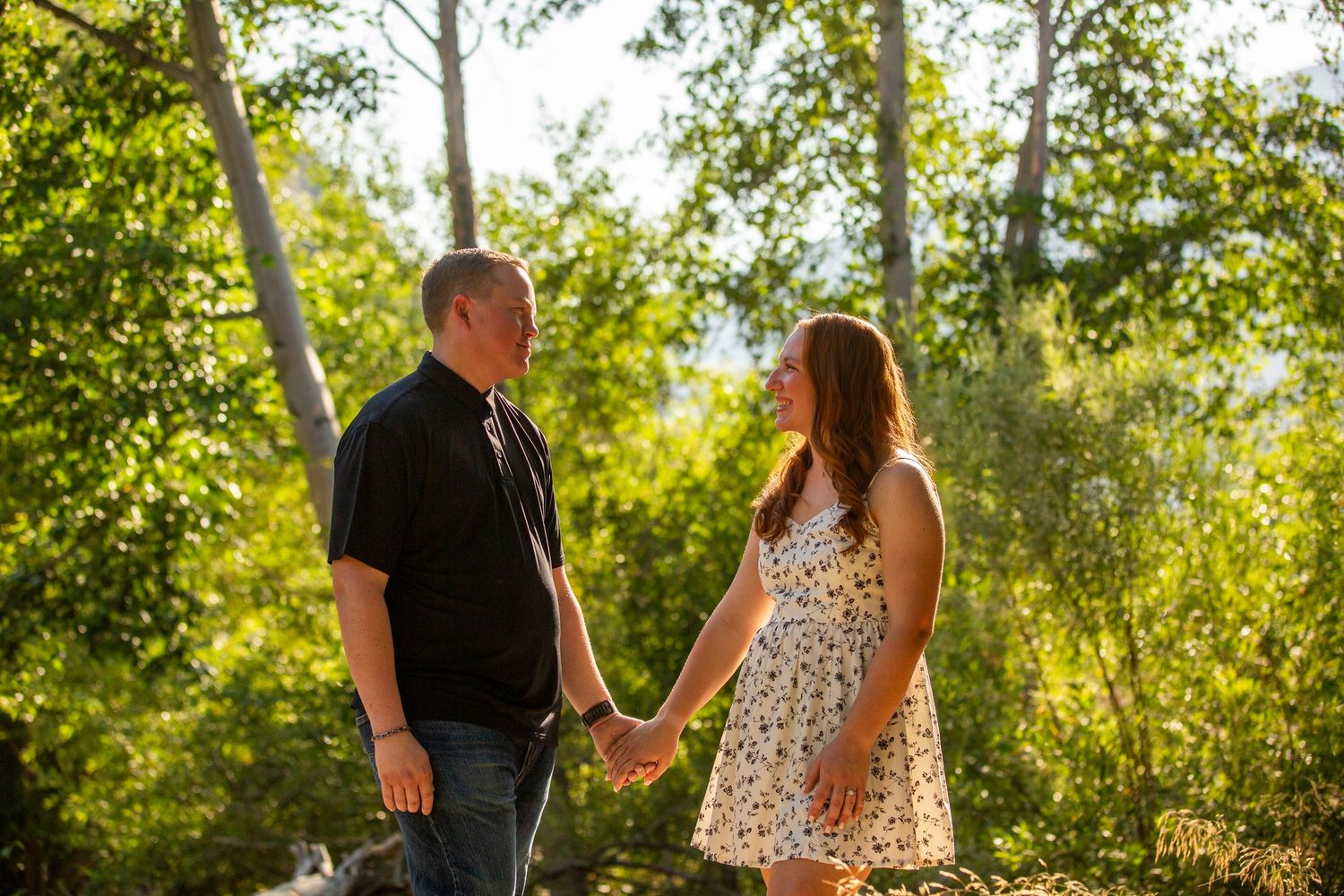 A couple walks together on a sunlit forest path during a summer afternoon, holding hands affectionately.