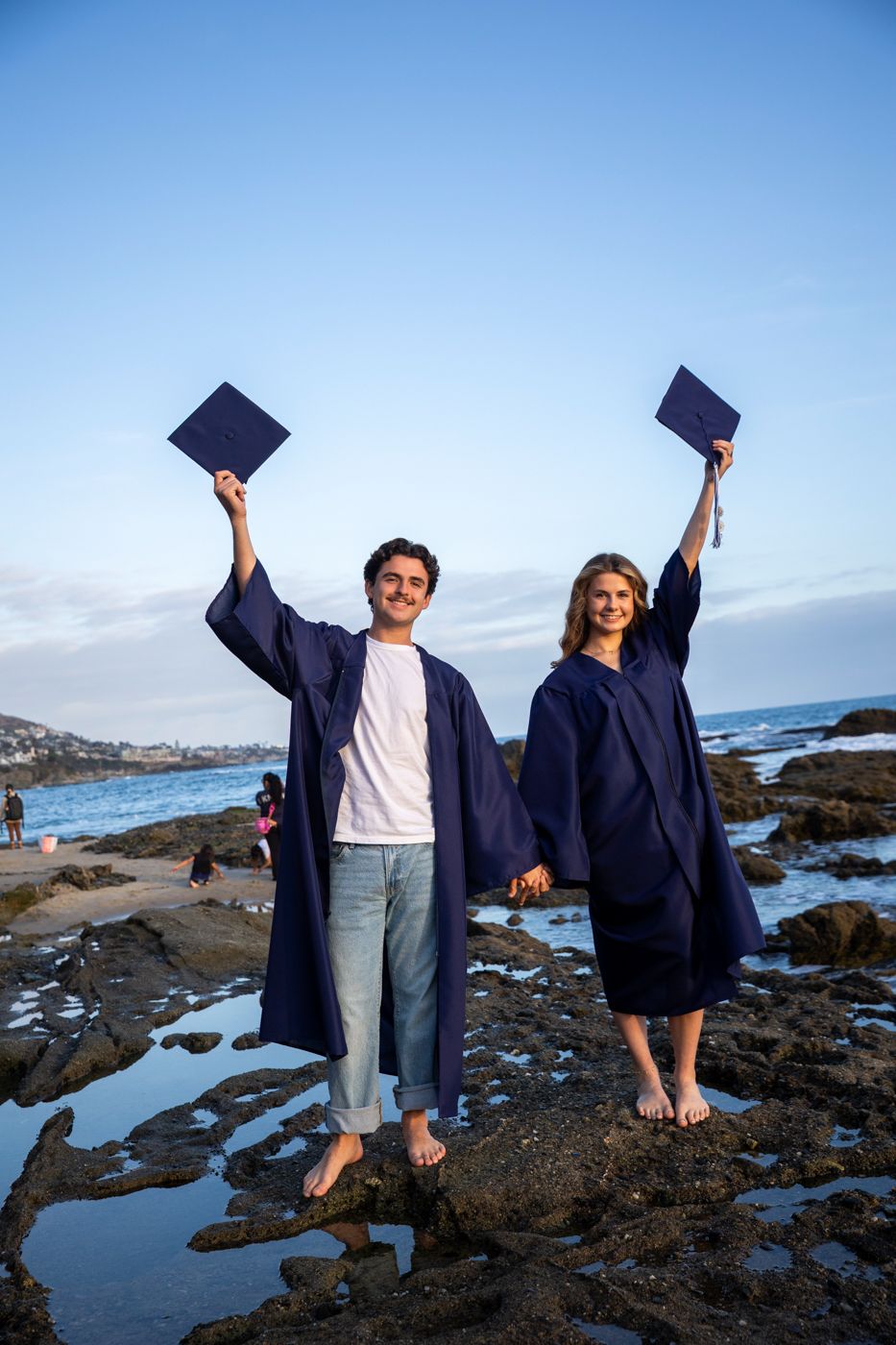 Graduates celebrate on rocky beach holding up diplomas against blue sky. Beach photography, Huntington Beach, Laguna Beach, Redlands photog