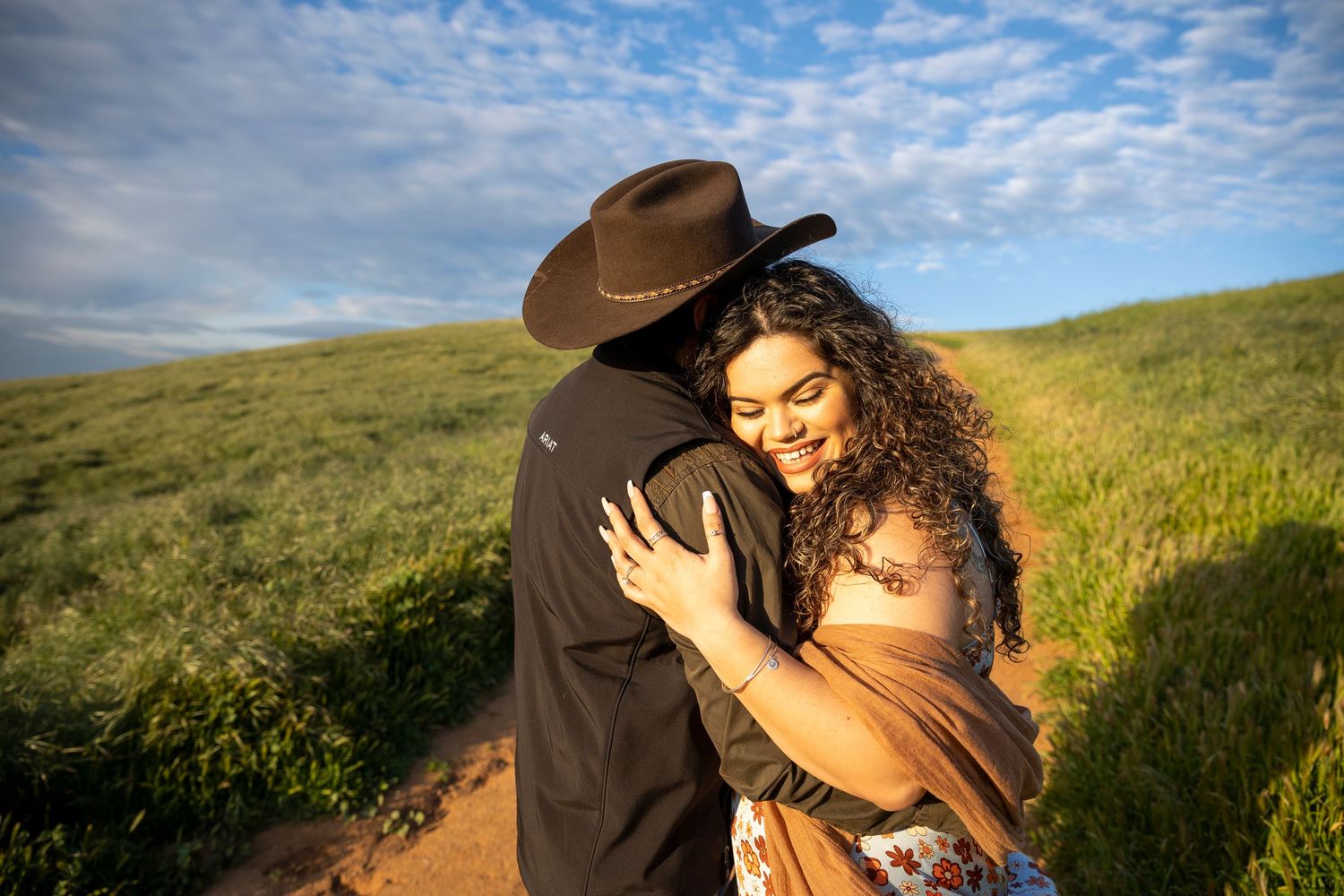 Couple embraces passionately in a sunlit grassy meadow with a cloudy sky backdrop at golden hour.