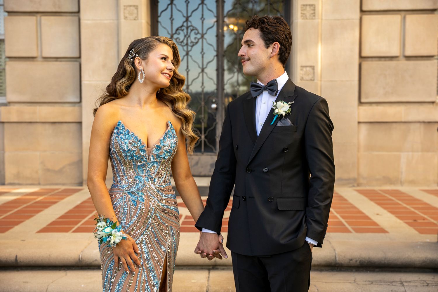 Prom Couple in formal attire smiling while holding hands outside historic building. San Bernardio photographer